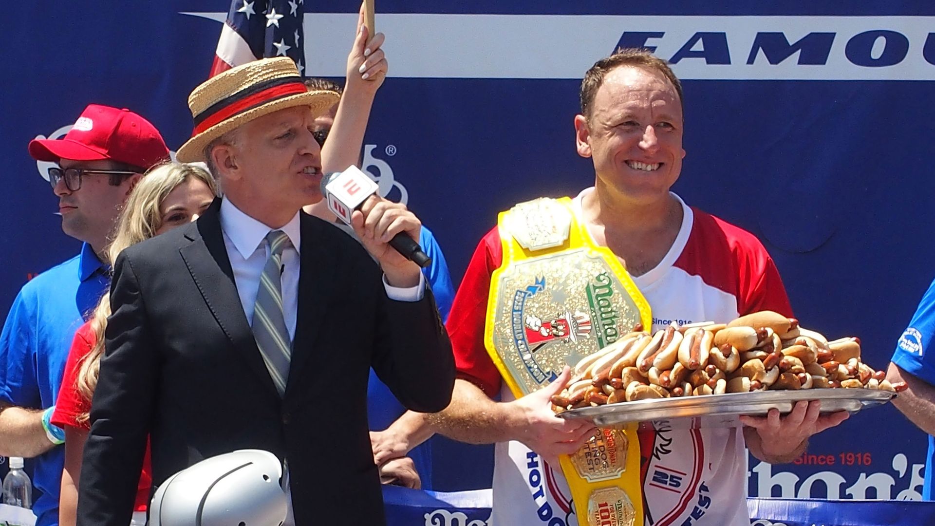 Joey Chestnut, wearing a red and white shirt wearing a championship belt, holds a tray piled with hot dogs, smiling. Another man in a suit and straw hat speaks into a microphone. Nathan's Hot Dog Contest.