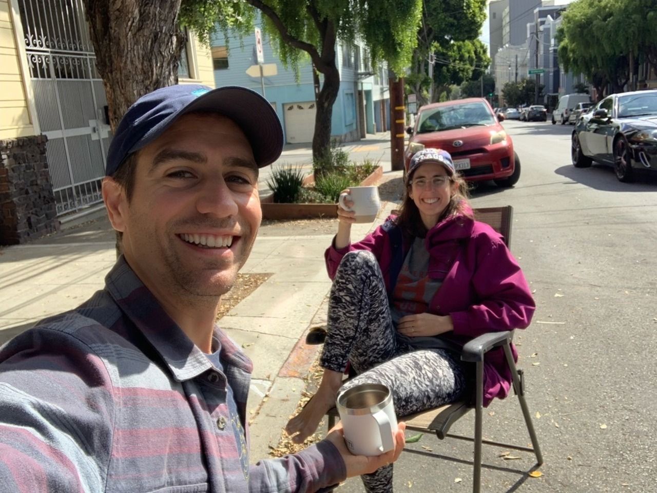 Man in a plaid shirt and navy cap takes a selfie with a woman in a magenta jacket and patterned leggings sitting barefoot on a chair on a sunny sidewalk, both holding white mugs.