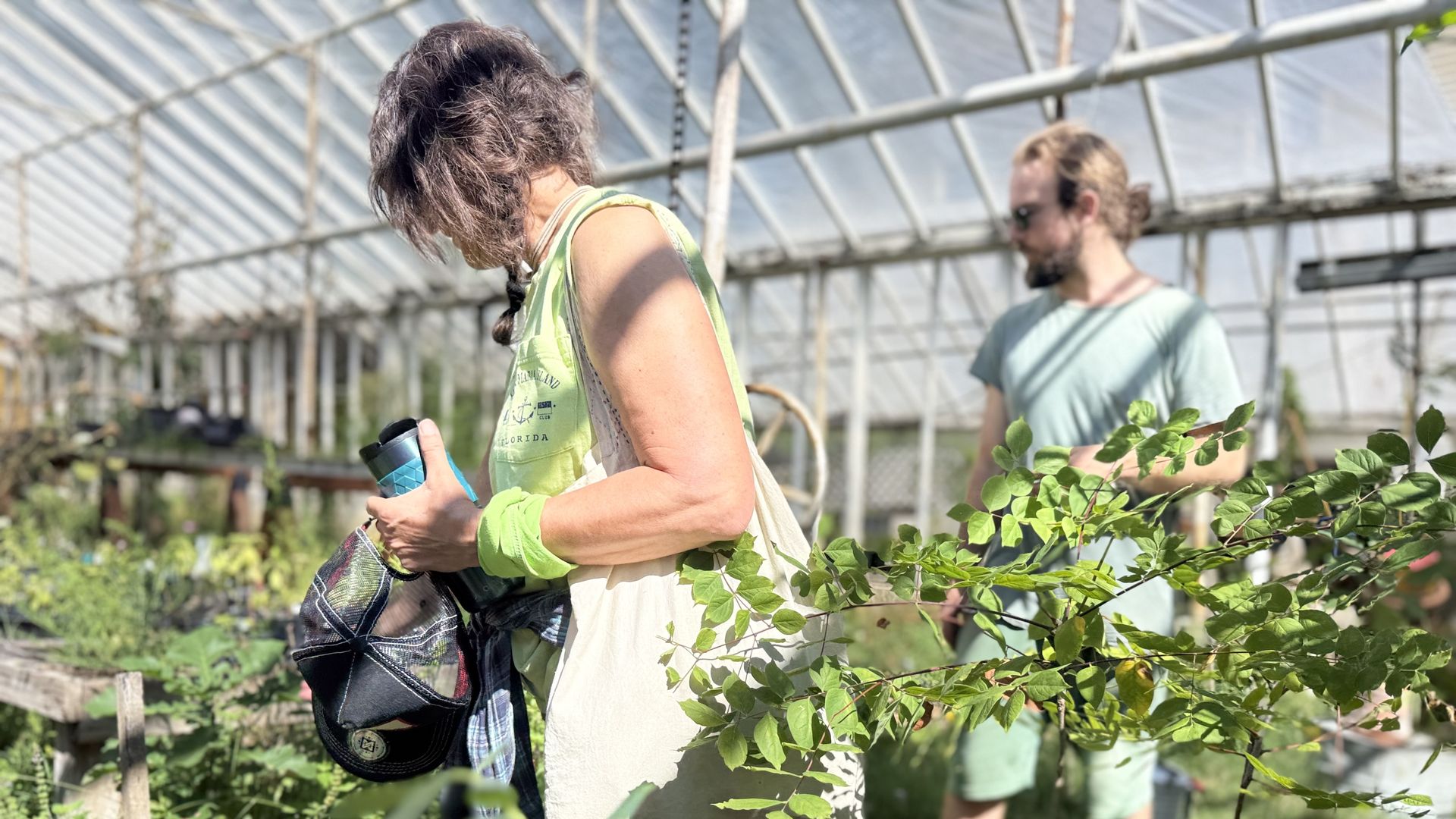 Two people in a bright greenhouse: a woman in a lime top carries a bag and a bottle among leafy vines, while a man in a pale teal shirt and sunglasses stands in the background.