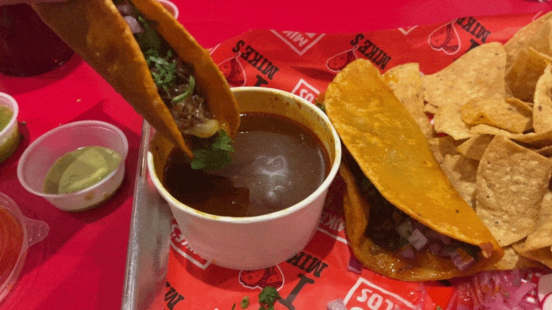 A hand dunks a beef birra taco into a red consome broth on a tray with another taco and tortilla chips. 