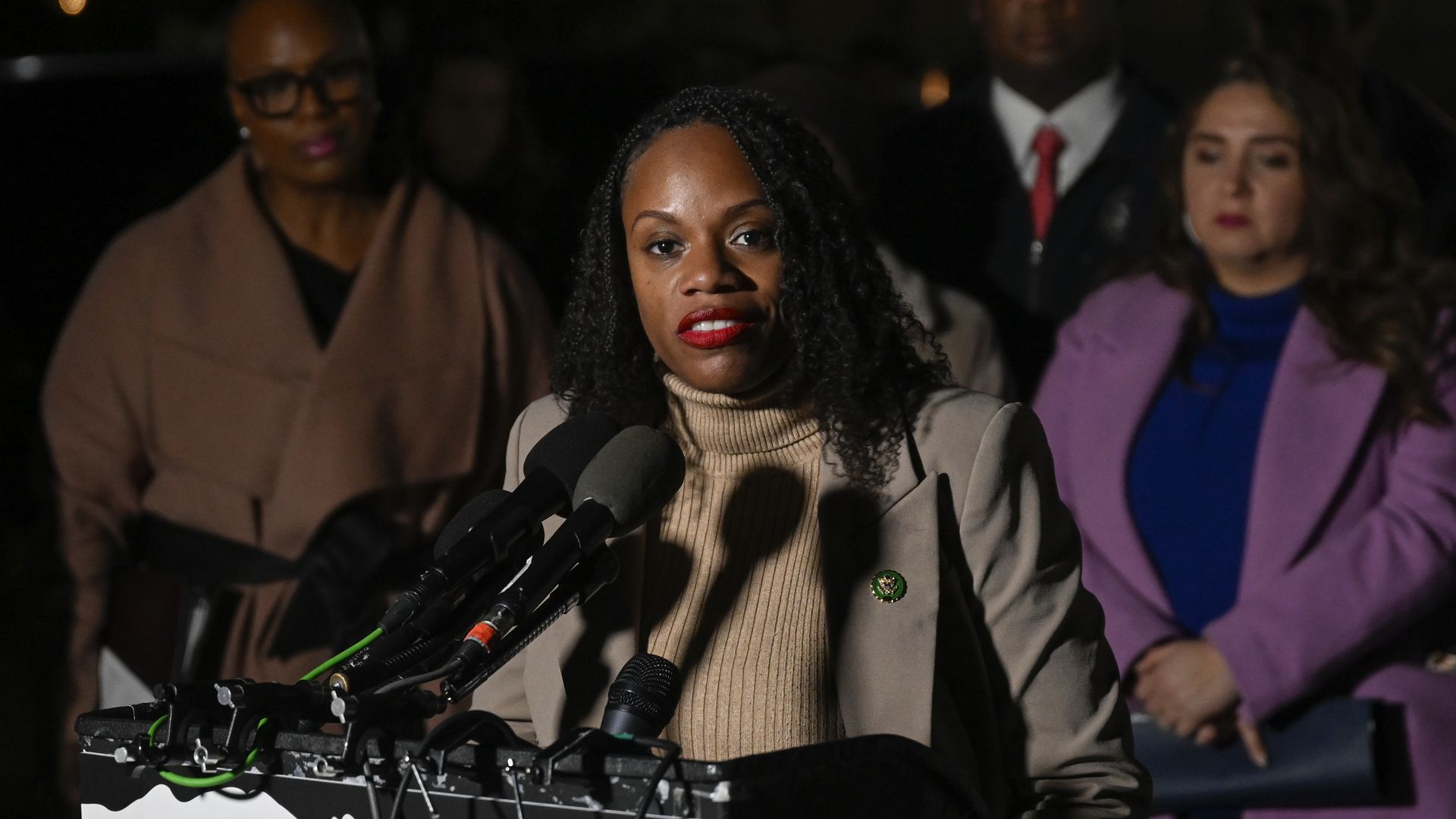 Rep. Summer Lee, wearing a tan overcoat and tan sweater, speaks at microphones surrounded by progressive colleagues.