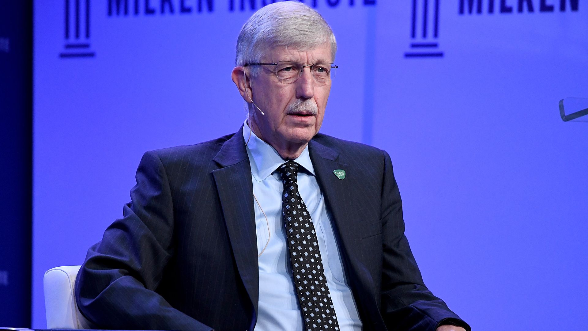 In this image, Collins is sitting in a chair wearing a suit.