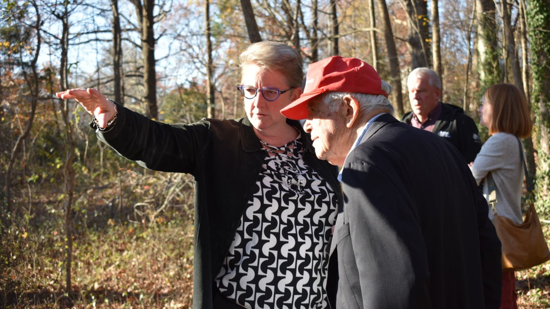 A woman and a man standing in the woods. The woman is pointing to something in the distance.
