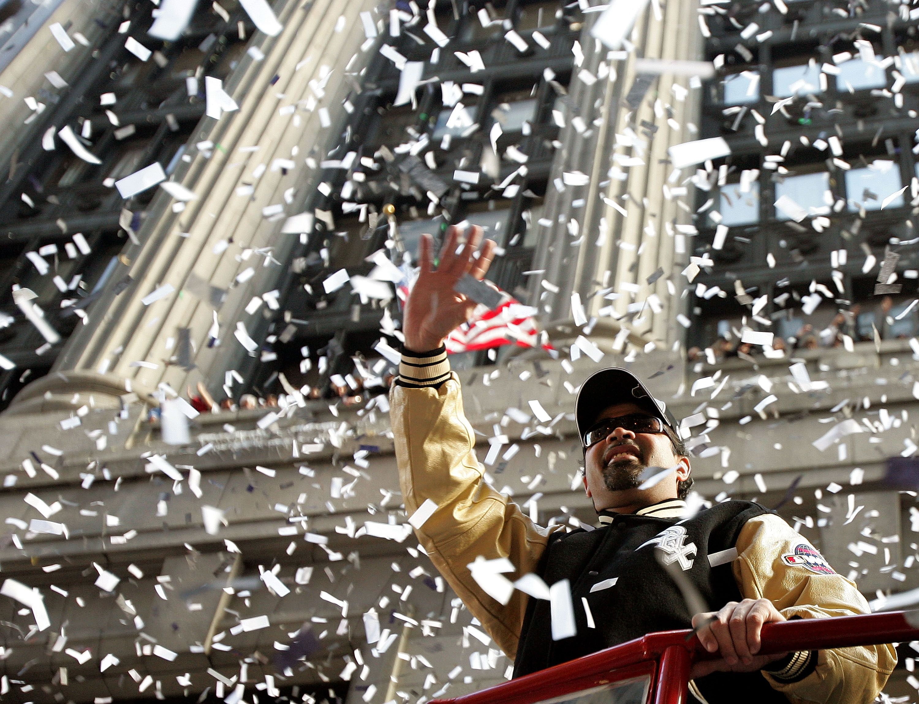 Two people ride a red float as white confetti rains down; a man in a beige jacket and black cap waves, wearing sunglasses, with a city backdrop.