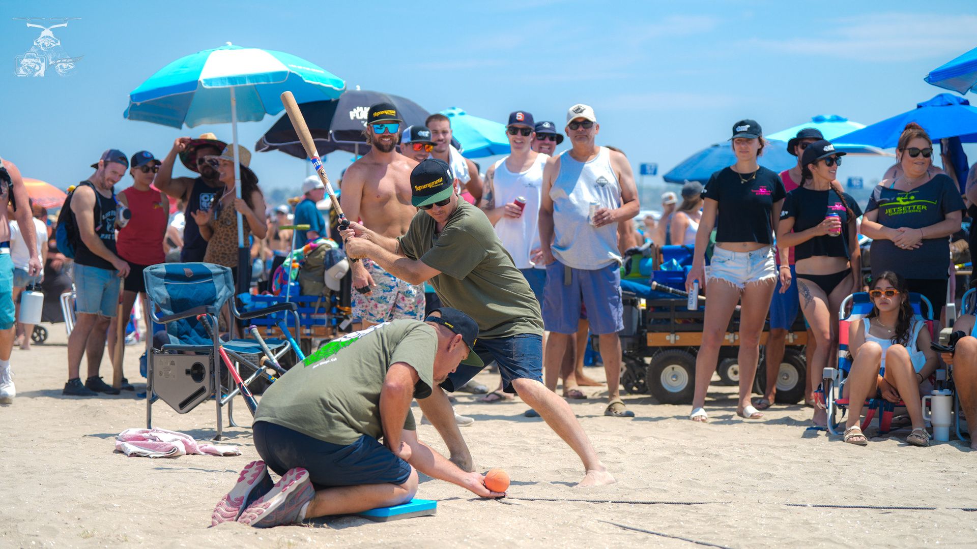 A crowd of people in beach wear watch two men in green tee shirts playing a wiffle-ball like game on the beach. 