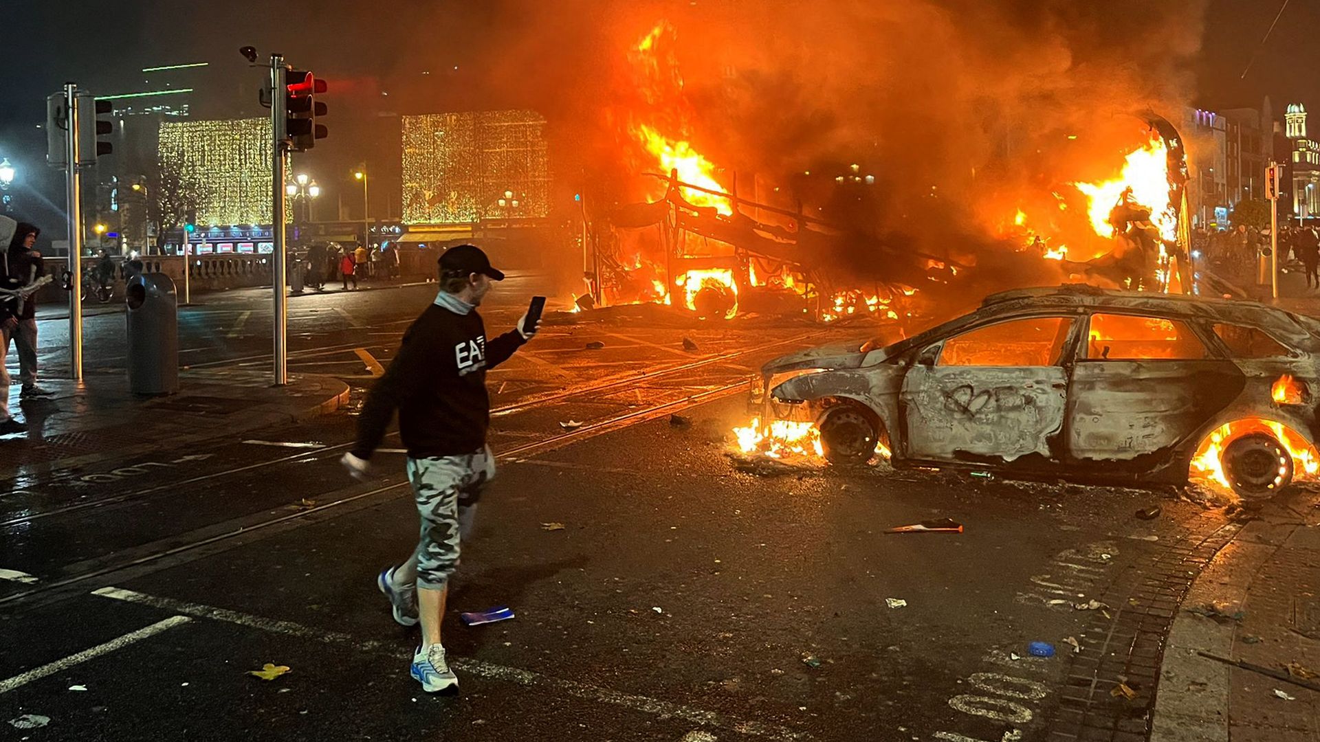 Fire engulfs the scene in the background in Dublin, with a car on fire and a person walking by on their phone in the foreground. 