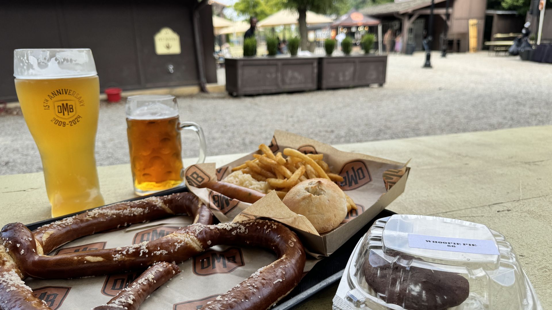 Outdoor beer garden scene with a tall pint and a smaller mug in the foreground, plus pretzels, fries, and a sandwich. Background shows blurred buildings, umbrellas, and trees.