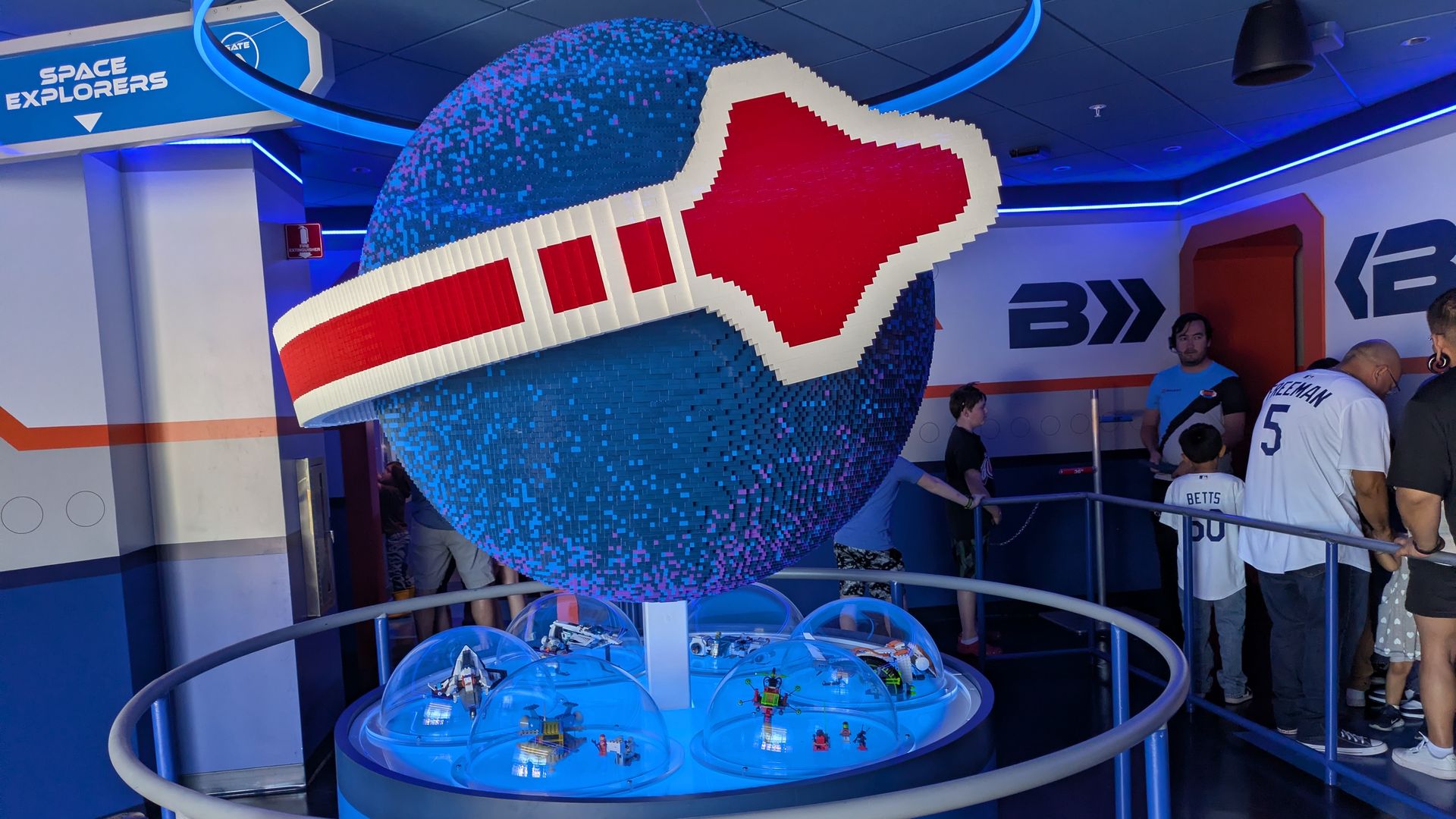 Blue pixelated globe on a pedestal with a white and red strap, ringed by a circular railing. Glass domes at the base house tiny figures in a space exhibit, with a "Space Explorers" sign and visitors nearby.