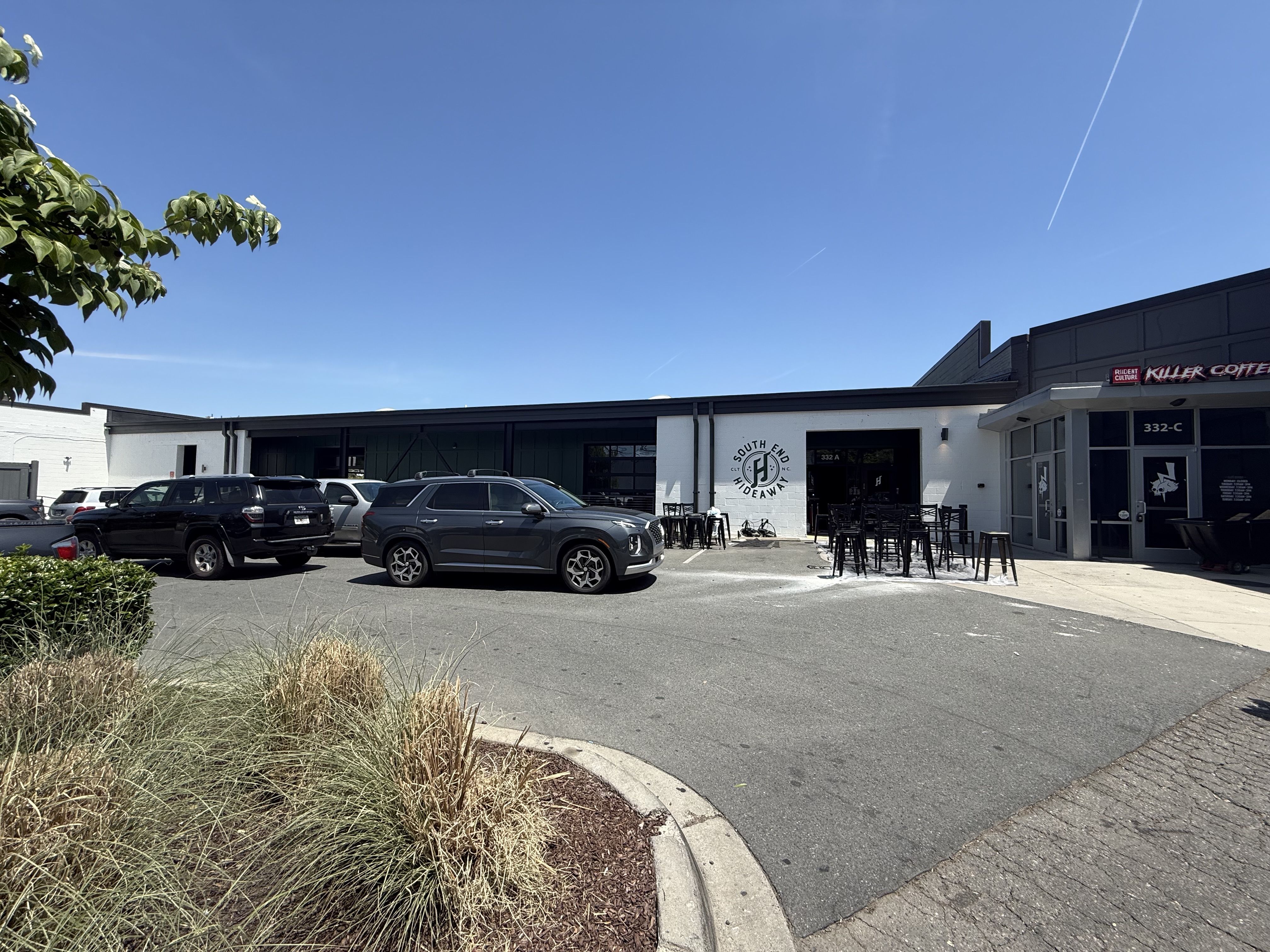 Blue sky with jet contrails over a strip mall. Foreground grasses, a parking lot full of cars, and a white building with outdoor seating. Sign reads "South End Hideaway" near the entrance; "Killer Coffee" on the right.