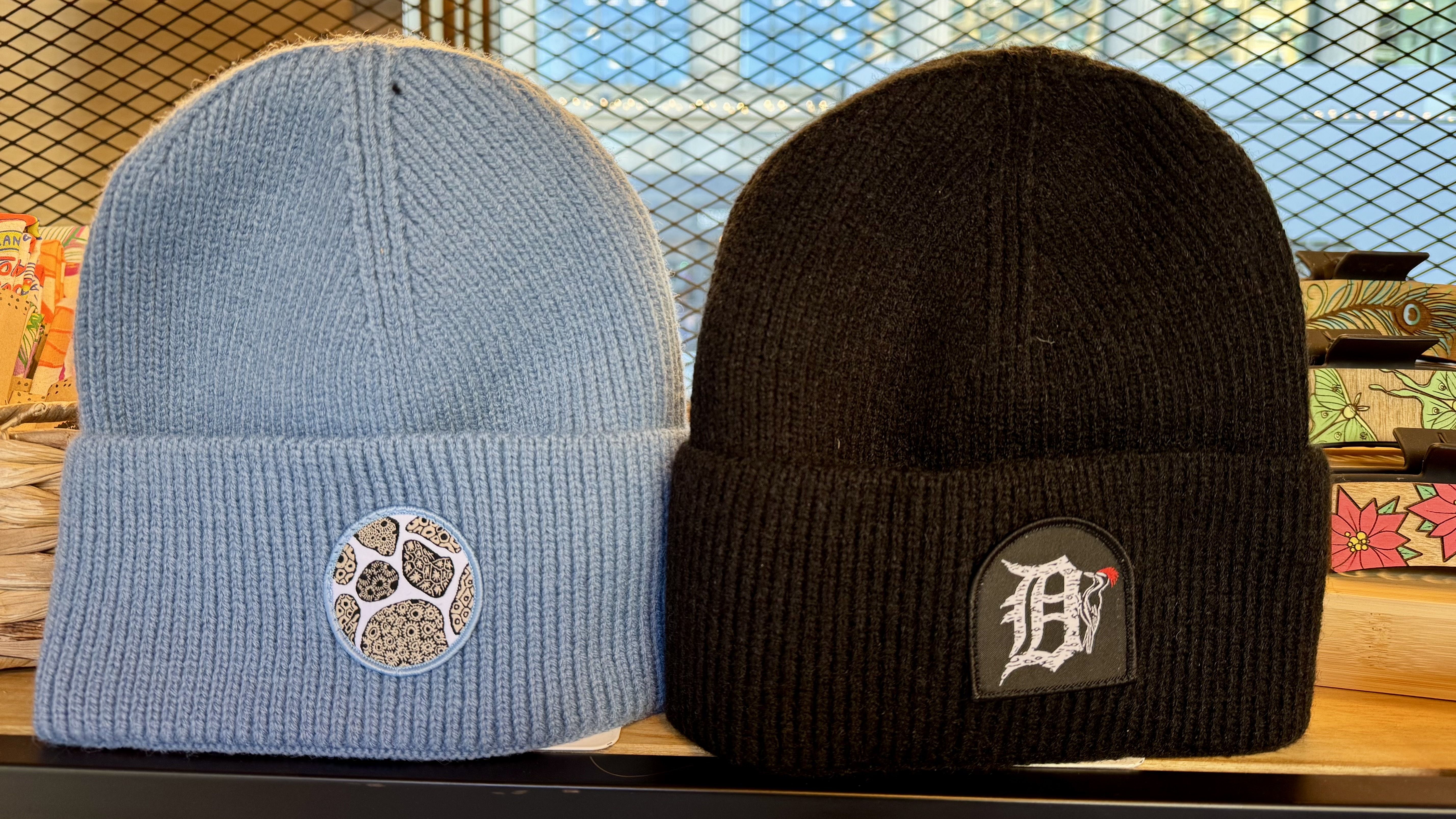 Two beanies, one light blue and one black. One features art of several petoskey stones and the other an old english D