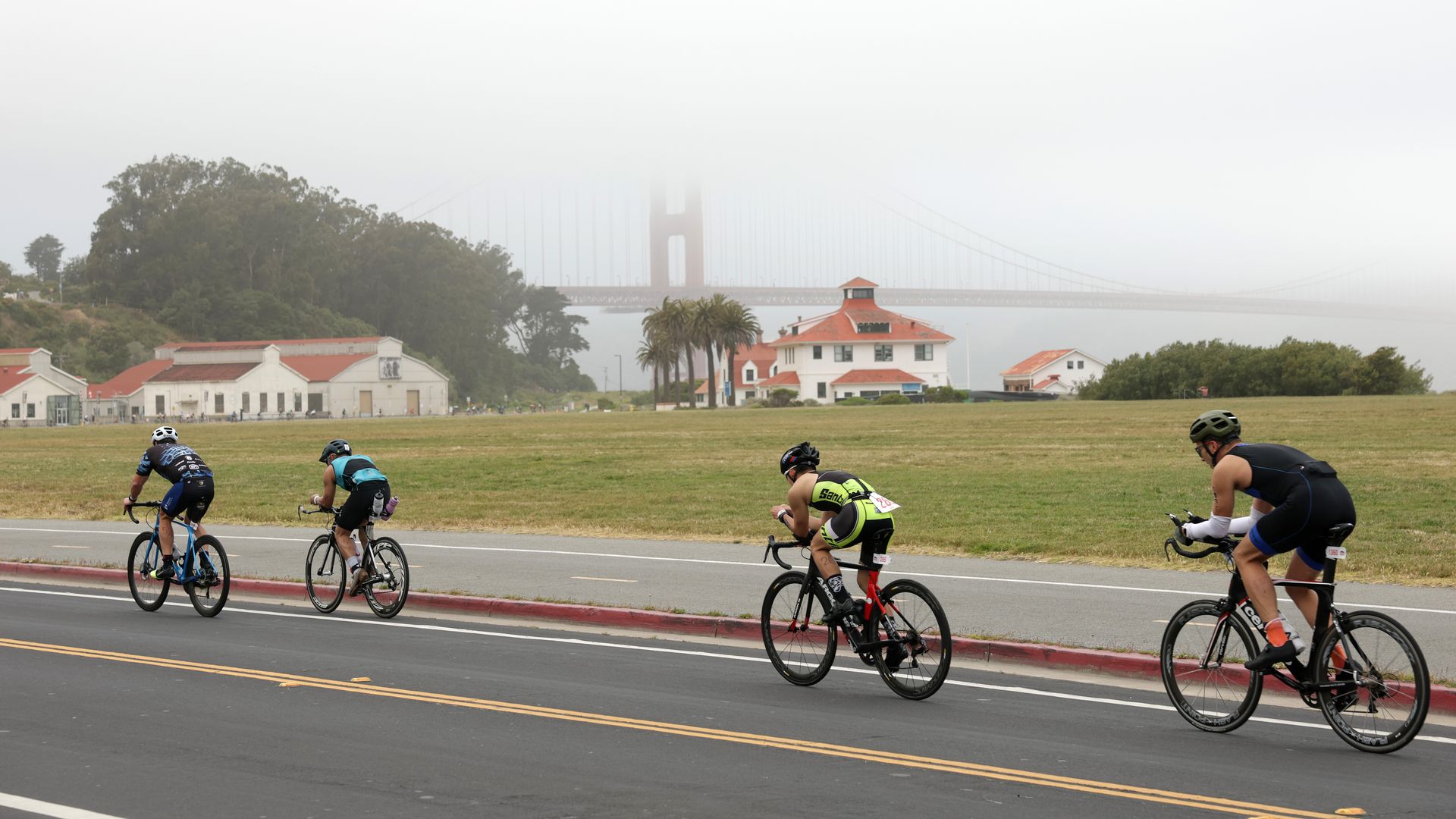 Four cyclists riding on a road near green fields with several white buildings having red roofs; fog partially obscures the iconic Golden Gate Bridge in the background.