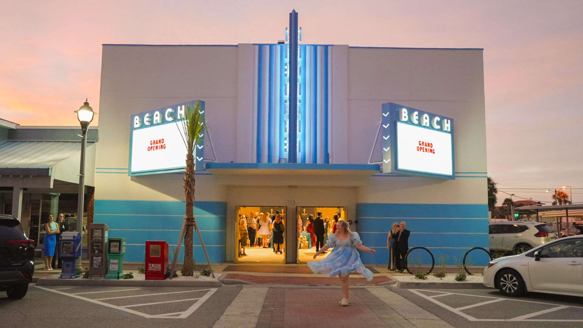 A woman in a cloud-print dress twirls on a brick crosswalk in front of a white-and-blue art deco building with marquees that say "BEACH" in white letters on top and "GRAND OPENING" in red letters in the message boards.