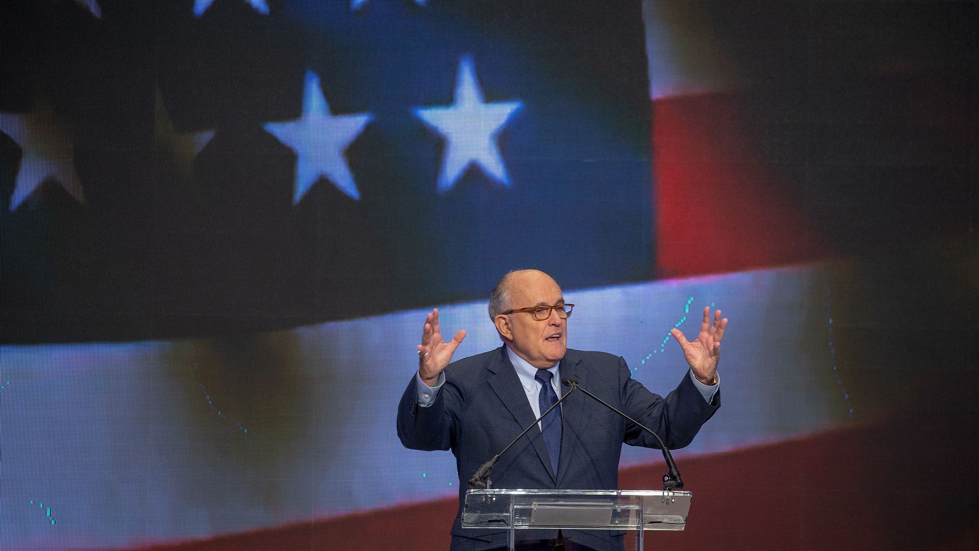 Rudy Giuliani lifts his hands while speaking at a podium. An American flag is shown on a screen behind him
