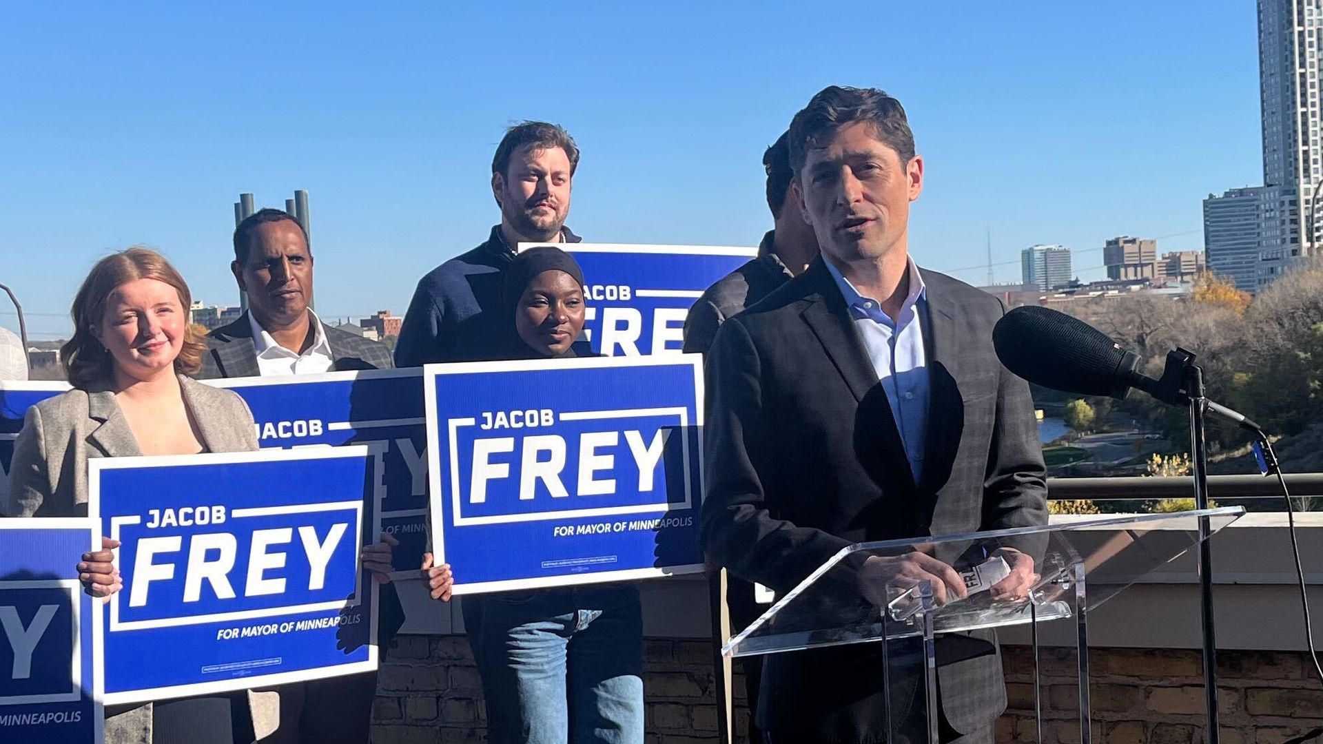 A man in a dark plaid suit speaks at a clear podium with a microphone on a rooftop, surrounded by six people holding blue signs reading "Jacob Frey for Mayor of Minneapolis," under a clear blue sky.
