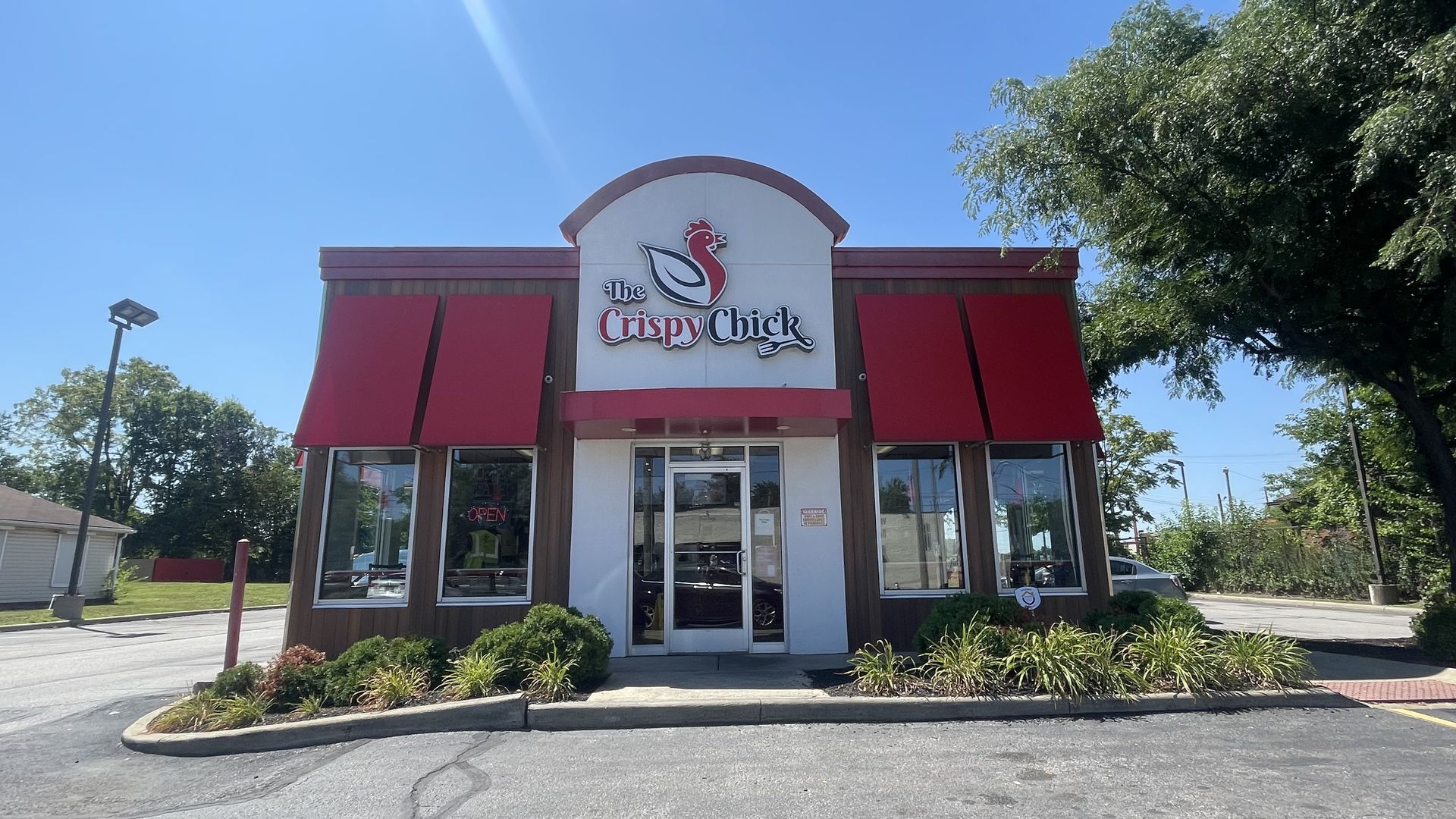 Front view of a fast-food restaurant featuring the sign "The Crispy Chick" with a red chicken logo, white facade, red trim, glass windows, and green shrubs under a clear blue sky.