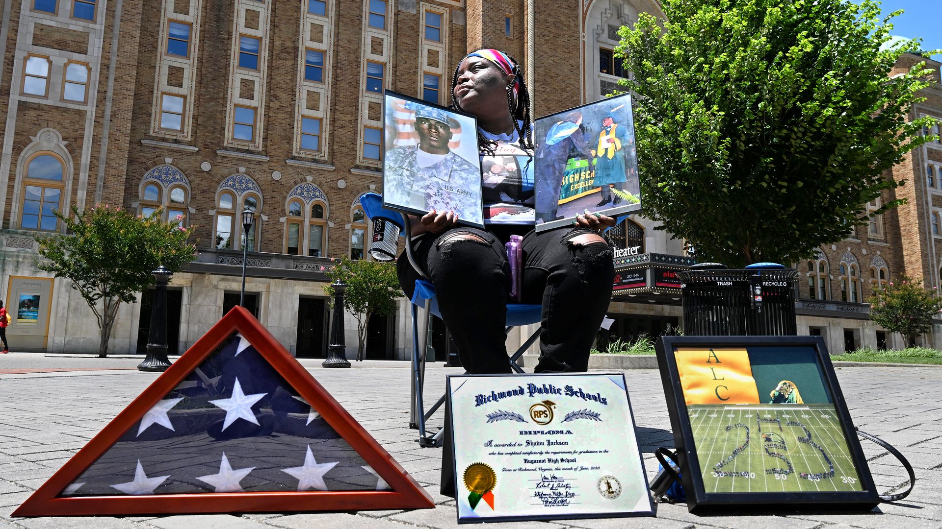 A woman sitting in front of the Altria Theater looking off to the left while holding photos of her husband and son in front of a diploma
