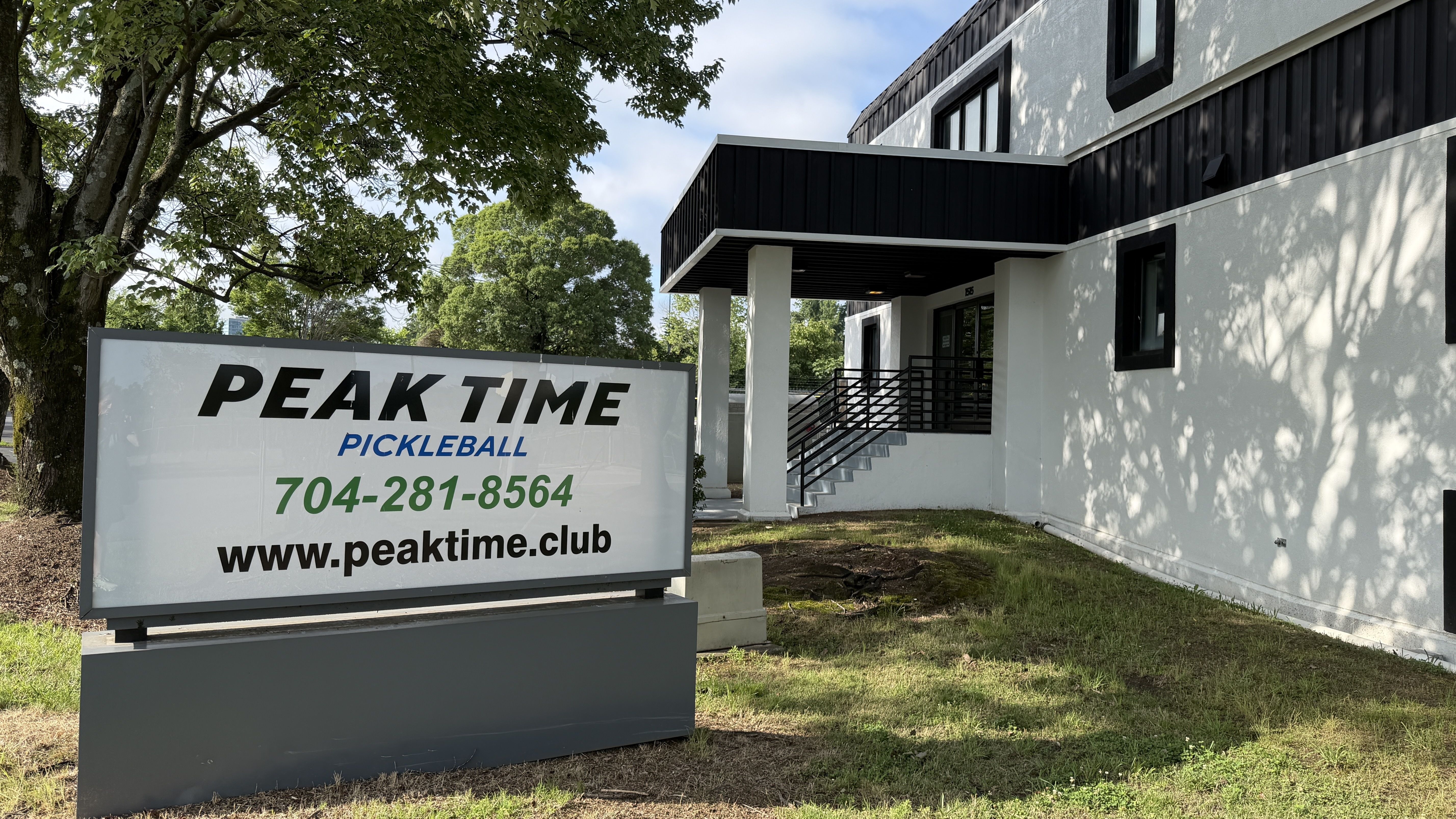 Modern white building with black trim and a large sign: "PEAK TIME" in black, "PICKLEBALL" in blue; green phone number and website visible. Trees and blue sky frame the scene.