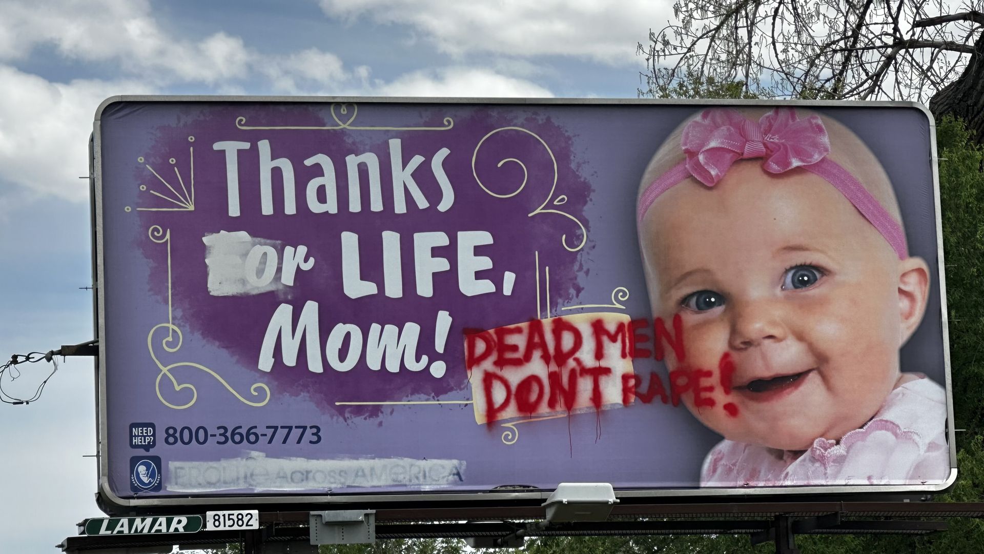Blue sky with white clouds above a large purple billboard showing a baby with a pink headband on the right; white decorative text about life on the left, red sticker partially covering it, helpline 800-366-7773.