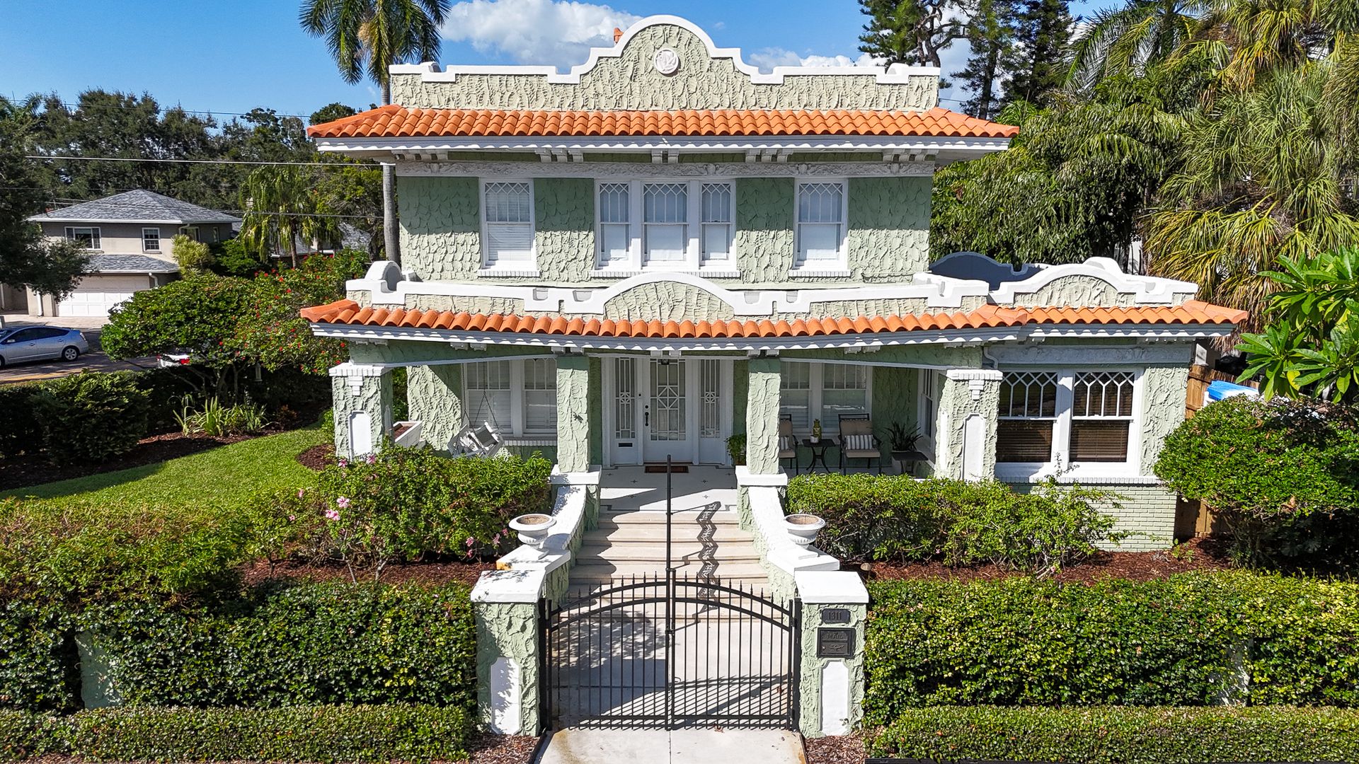 Front view of a two-story green house with textured walls, white trim, and red tiled roof, featuring a black metal gate, manicured bushes, and a porch with chairs under clear blue sky.