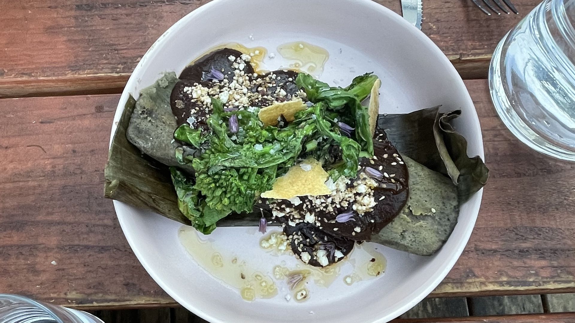 A photo of a blue corn tamale in a white plate on a wood-grain table. It's topped with green garnishes and white cheese.
