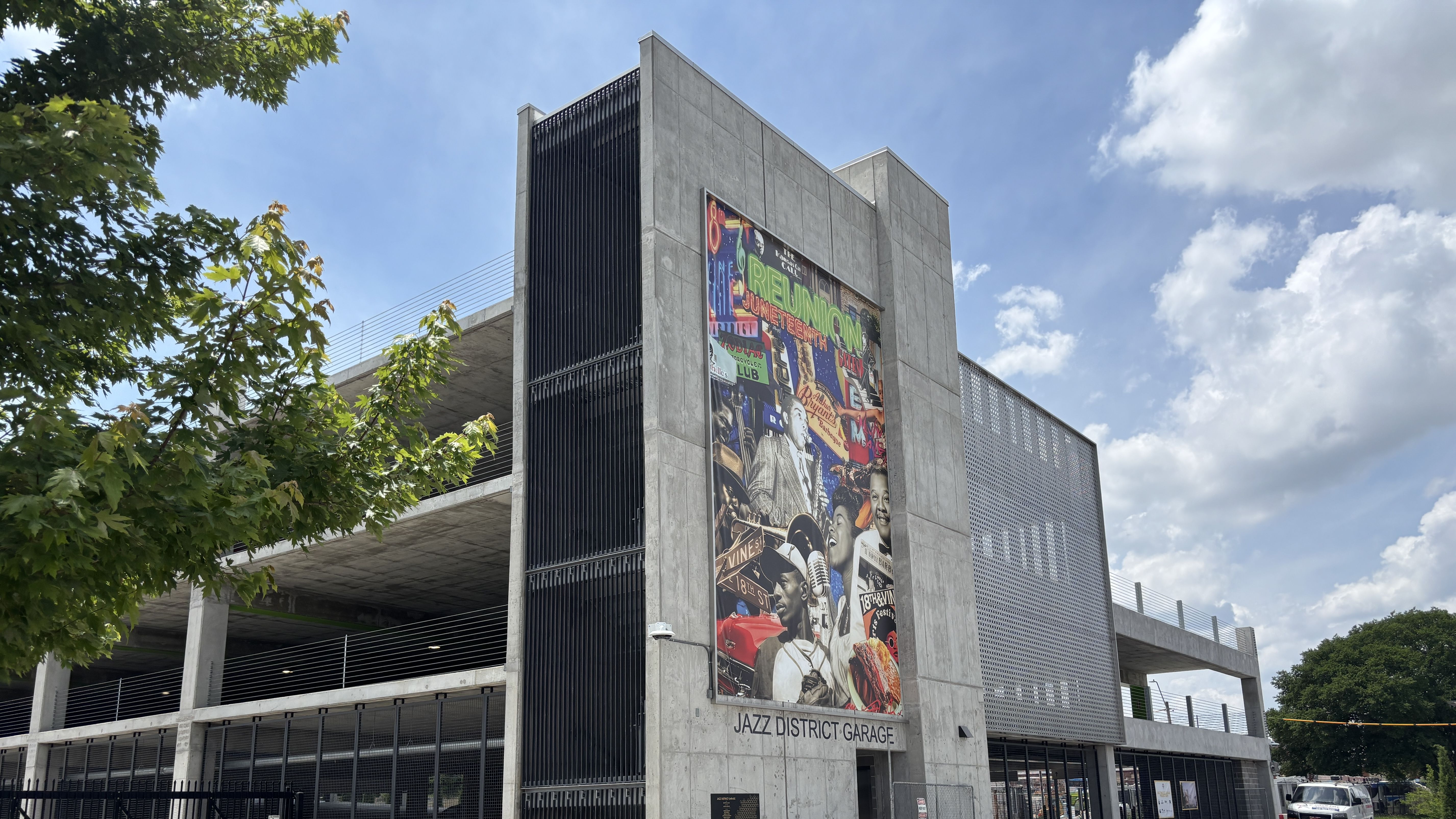 Concrete parking garage with a large colorful jazz mural on the front, sign reading "Jazz District Garage"; green tree on the left and a bright blue sky with clouds.