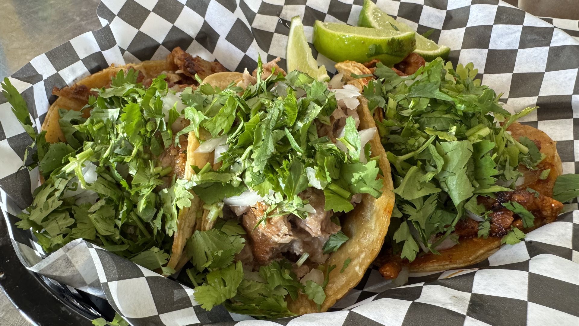 Two soft tacos filled with meat, onions, and a generous amount of fresh cilantro, served in a black-and-white checkered liner with lime wedges on the side.
