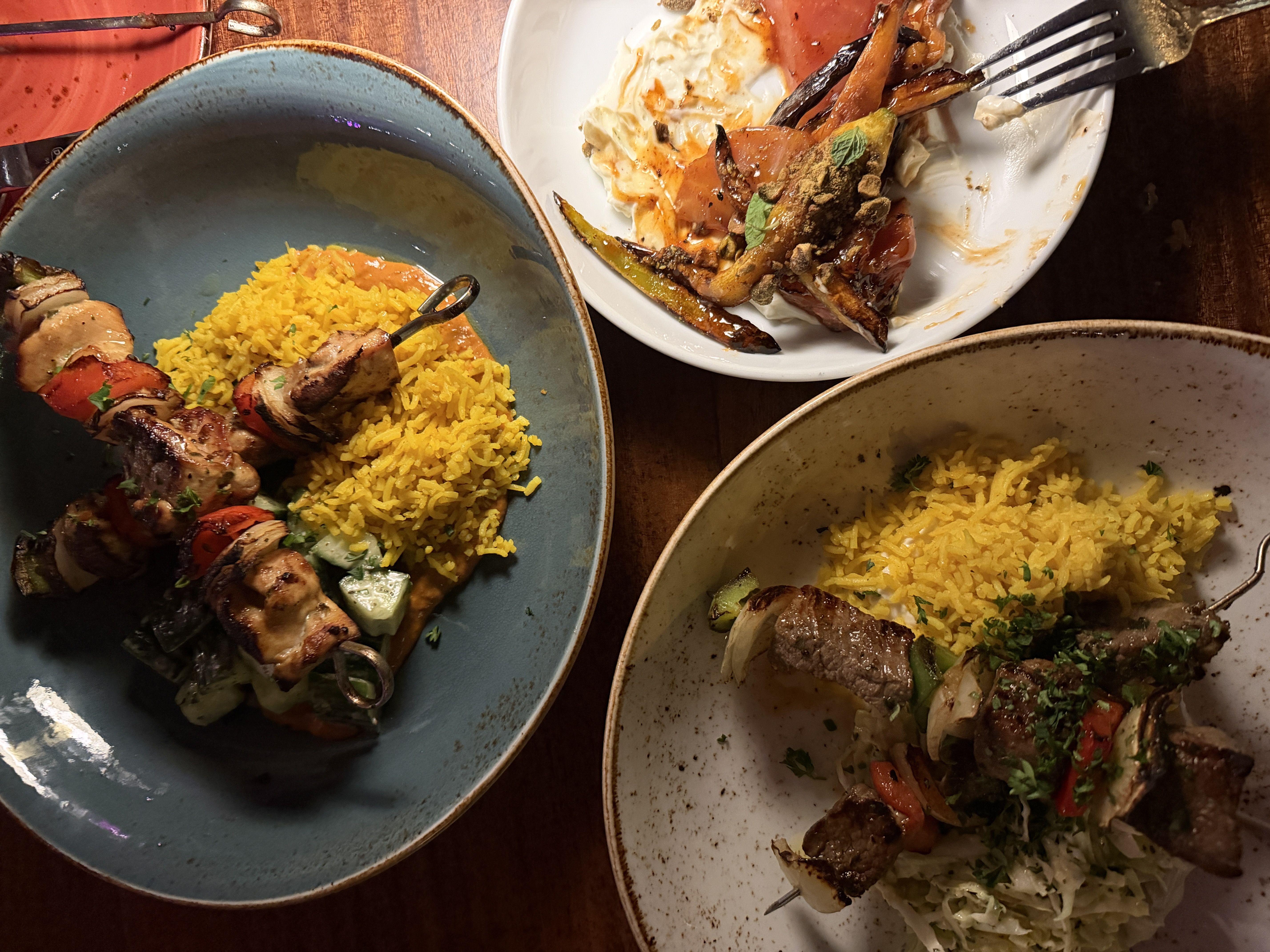 Two plates with skewers of grilled meat and vegetables, yellow rice, and salad; a third plate with roasted carrots, sauce, and a dollop of cream on a wooden table.