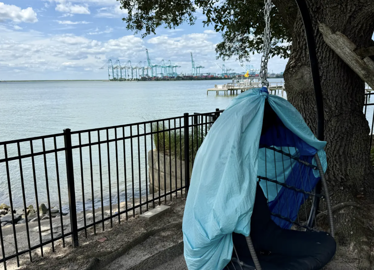 Blue and black hanging chair with fabric cover, positioned by a large tree and black metal fence overlooking calm water and a distant industrial port with cranes under a cloudy sky.