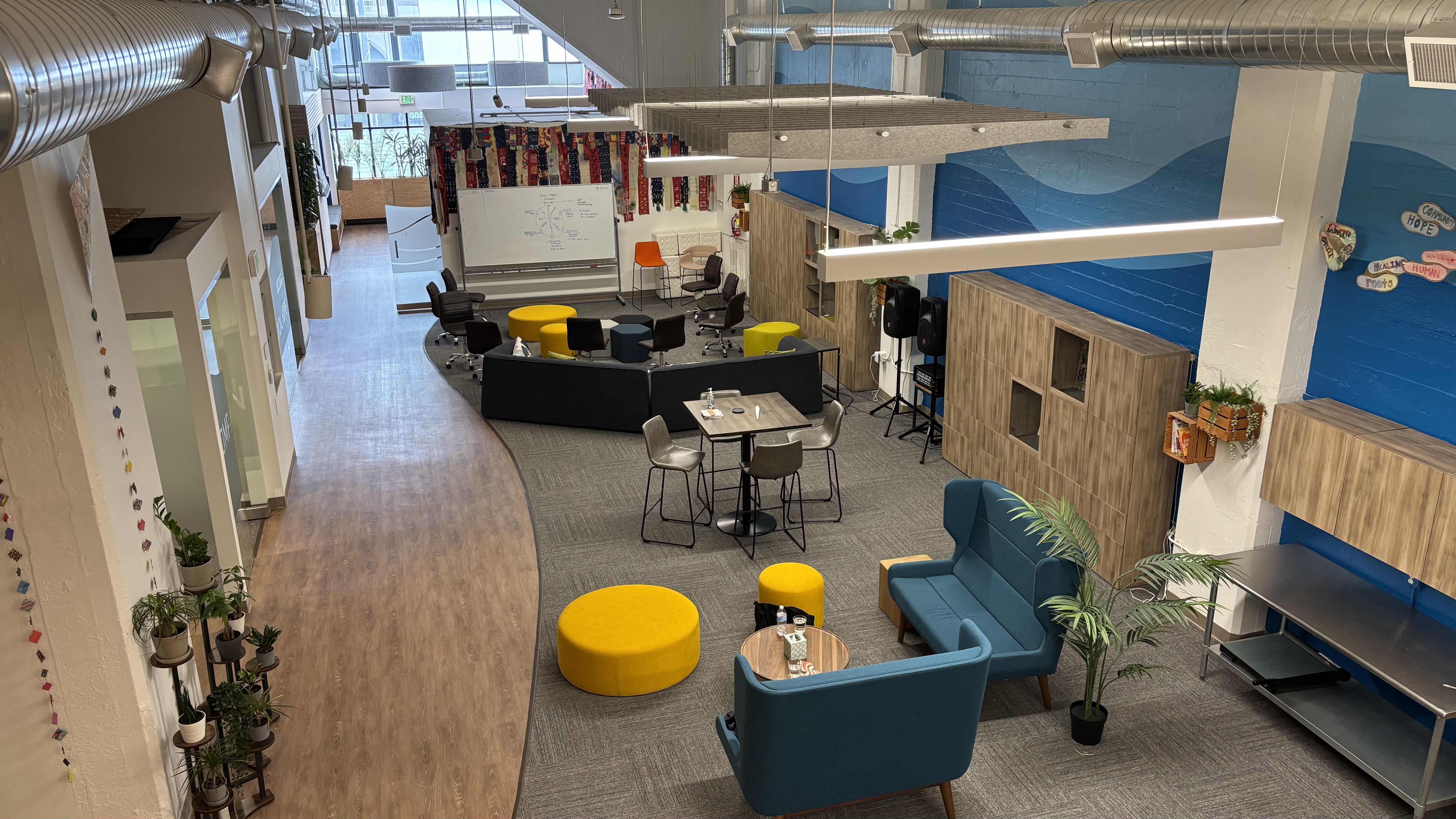 Modern office lounge with blue walls, wooden cabinets, blue sofas, yellow ottomans, black chairs, round tables, potted plants, and a whiteboard near large windows.