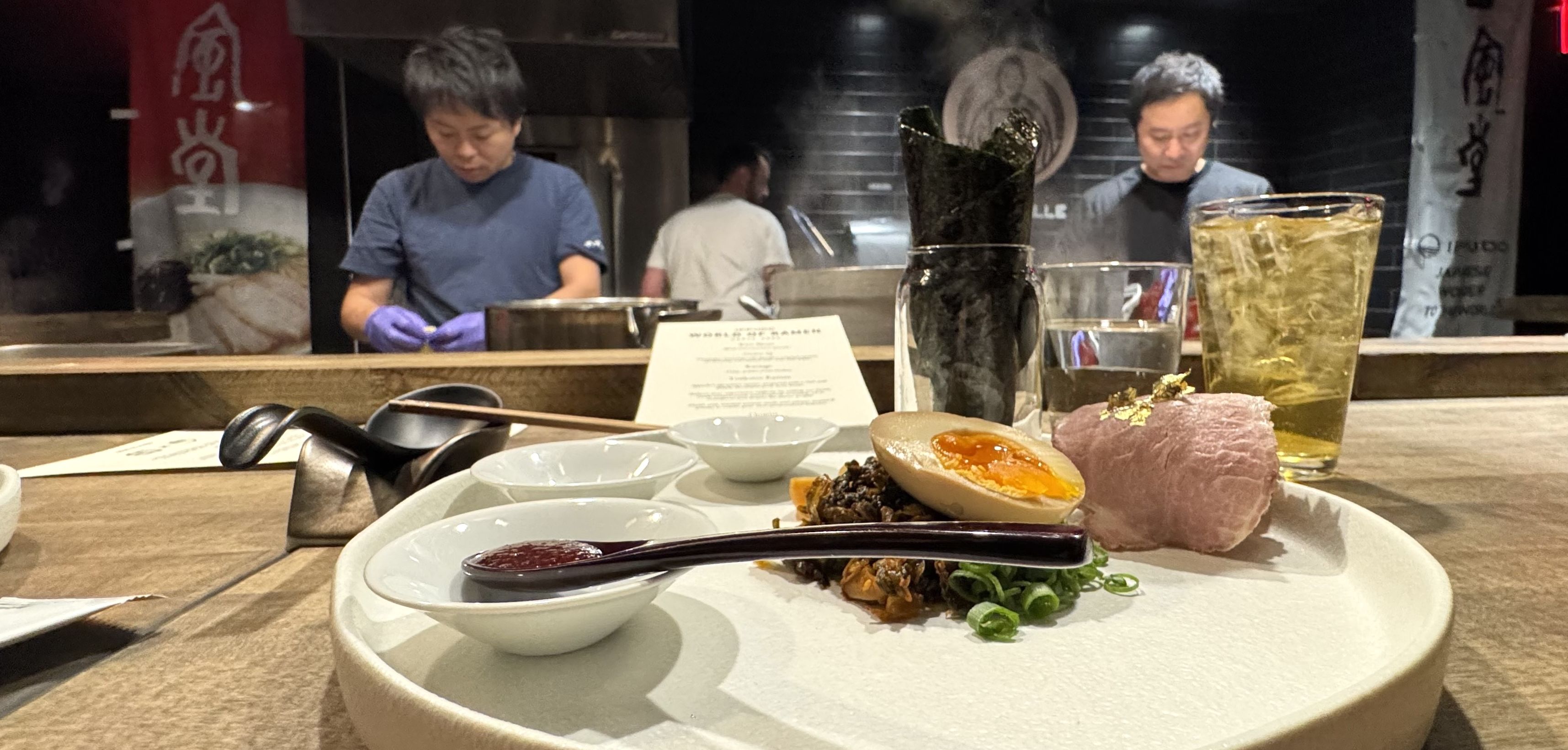 Close-up of a Japanese-inspired dish with marinated egg, sliced meat topped with gold flakes, seaweed, and sauces on a white plate, with chefs cooking in the background.