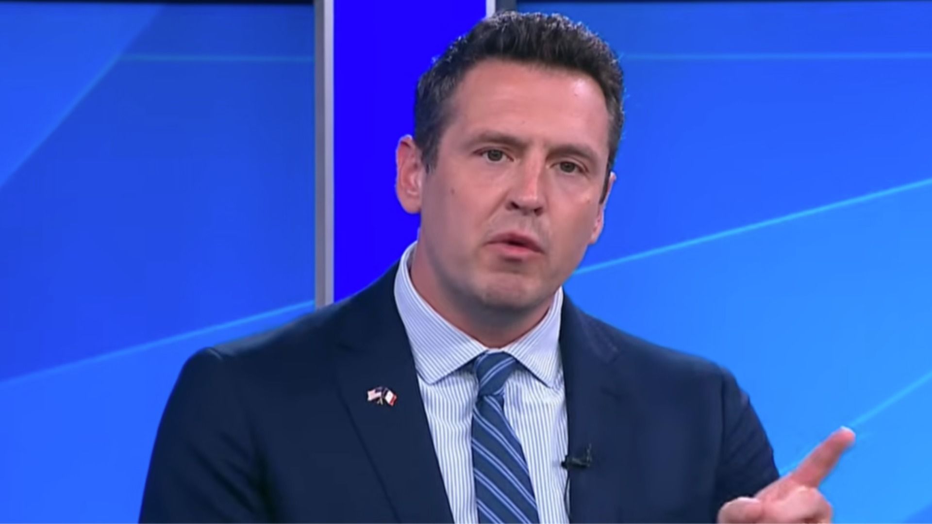 A man in a dark suit with a striped blue tie speaks on a blue TV studio set, gesturing with his right hand; a small flag lapel pin is visible.