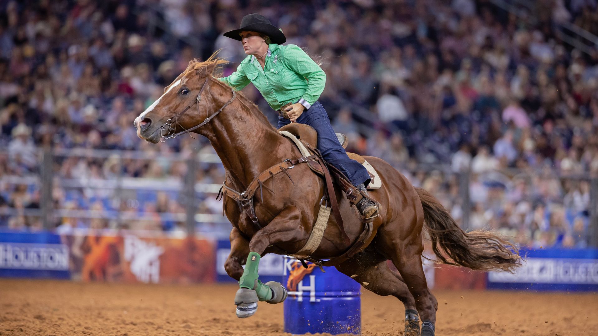 Rider in a green shirt and black hat on a galloping brown horse, jumping past a blue barrel in a crowded rodeo arena.