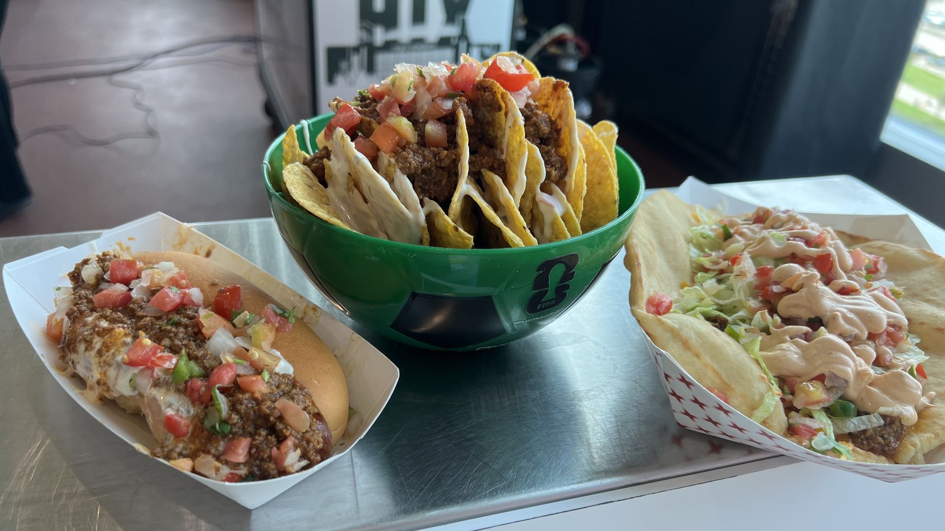 Green bowl of stacked hard-shell tacos filled with seasoned beef and pico de gallo, surrounded by other tacos and a hot dog with toppings on a metal counter.