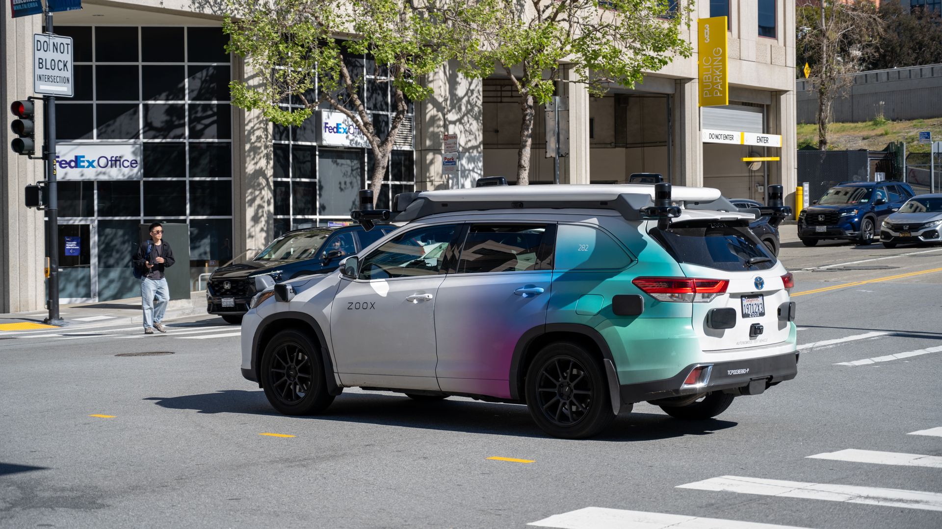 Photo of a white SUV with a colorful rear, with automotive mechanics on top, in an intersection. 