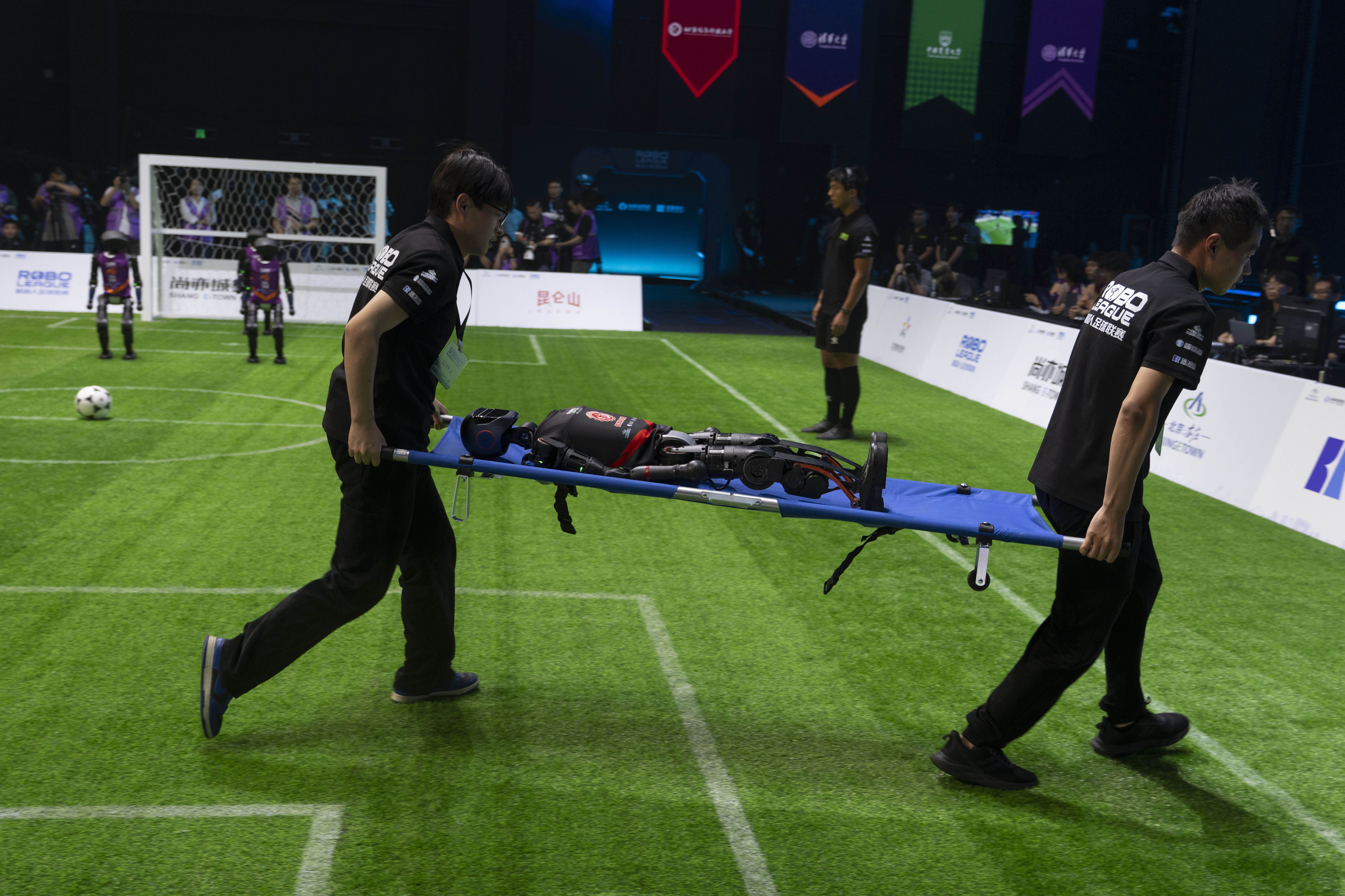 Two individuals in matching black uniforms carry a robot on a blue stretcher across a green artificial soccer field. The robot is humanoid and lies on its back with visible mechanical components. In the background, more robots can be seen on the field near a goal net, surrounded by spectators.