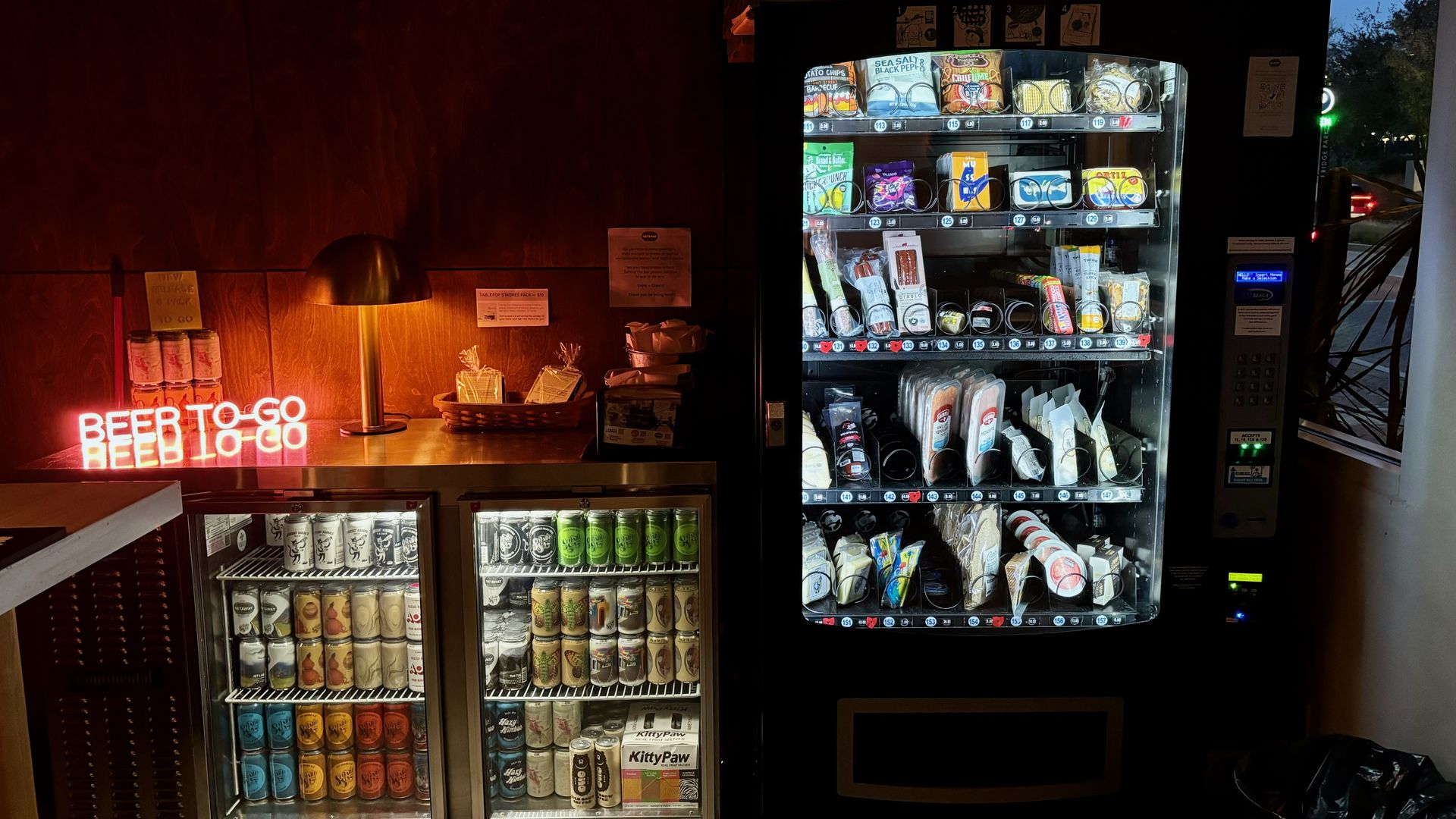A vending machine filled with snacks next to a fridge of beer cans and a neon sign that reads "beer to go"