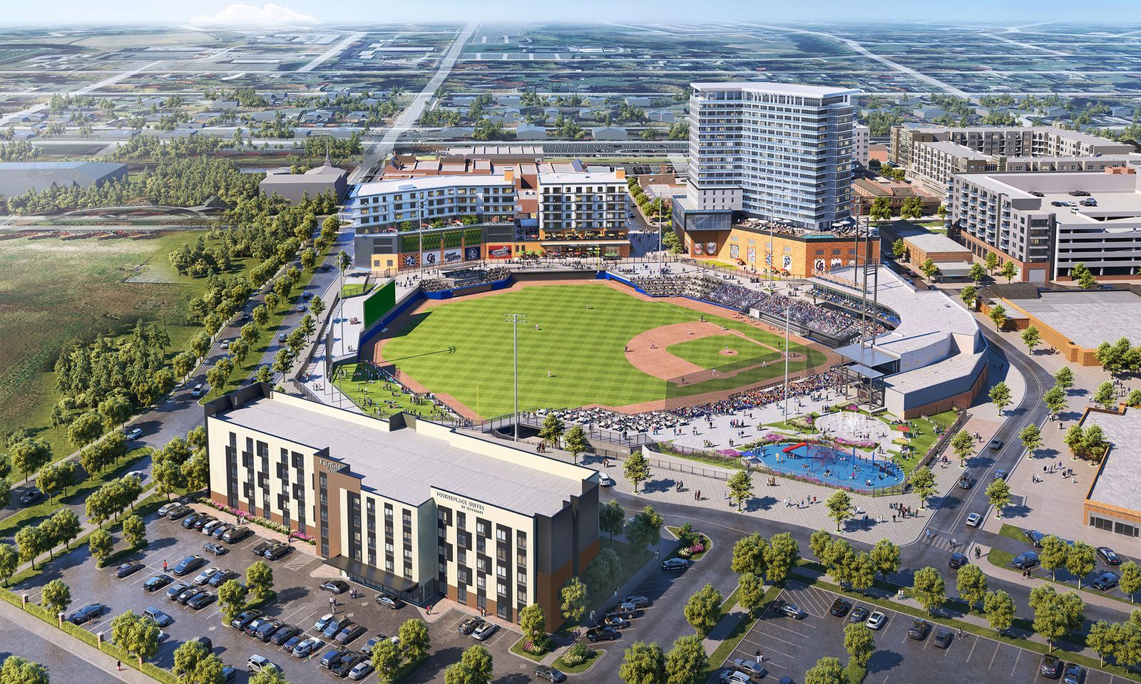 An aerial view of a modern baseball stadium in a mixed-use development: green field, surrounding stands, a curved shopping complex, a tall glass tower, parking lots, and tree-lined streets.