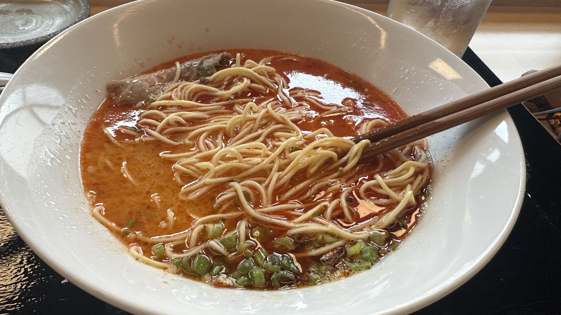White bowl of spicy ramen with red broth, tangled noodles, green onions, and pork slices. A pair of wooden chopsticks rests on the rim; glass and counter backdrop.