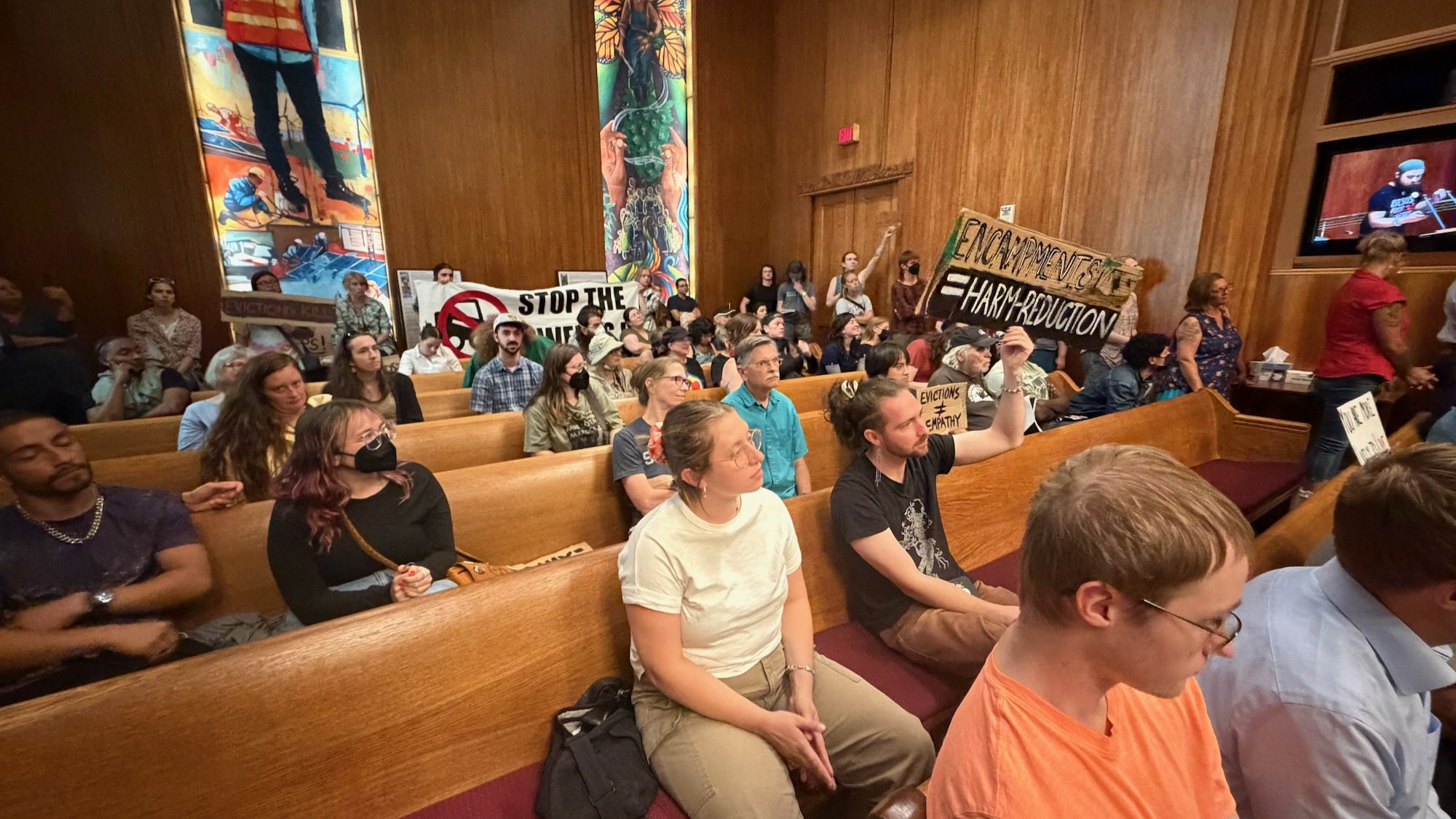A crowded church sanctuary with wooden walls and pews; protesters hold banners, including one reading "STOP THE" and another about harm reduction; attendees sit, some wearing masks.