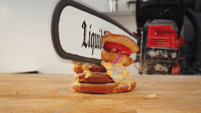 A sandwich with tomato, onion, and melted cheese is being sliced in half by a large chainsaw blade on a wooden table with sawdust around.