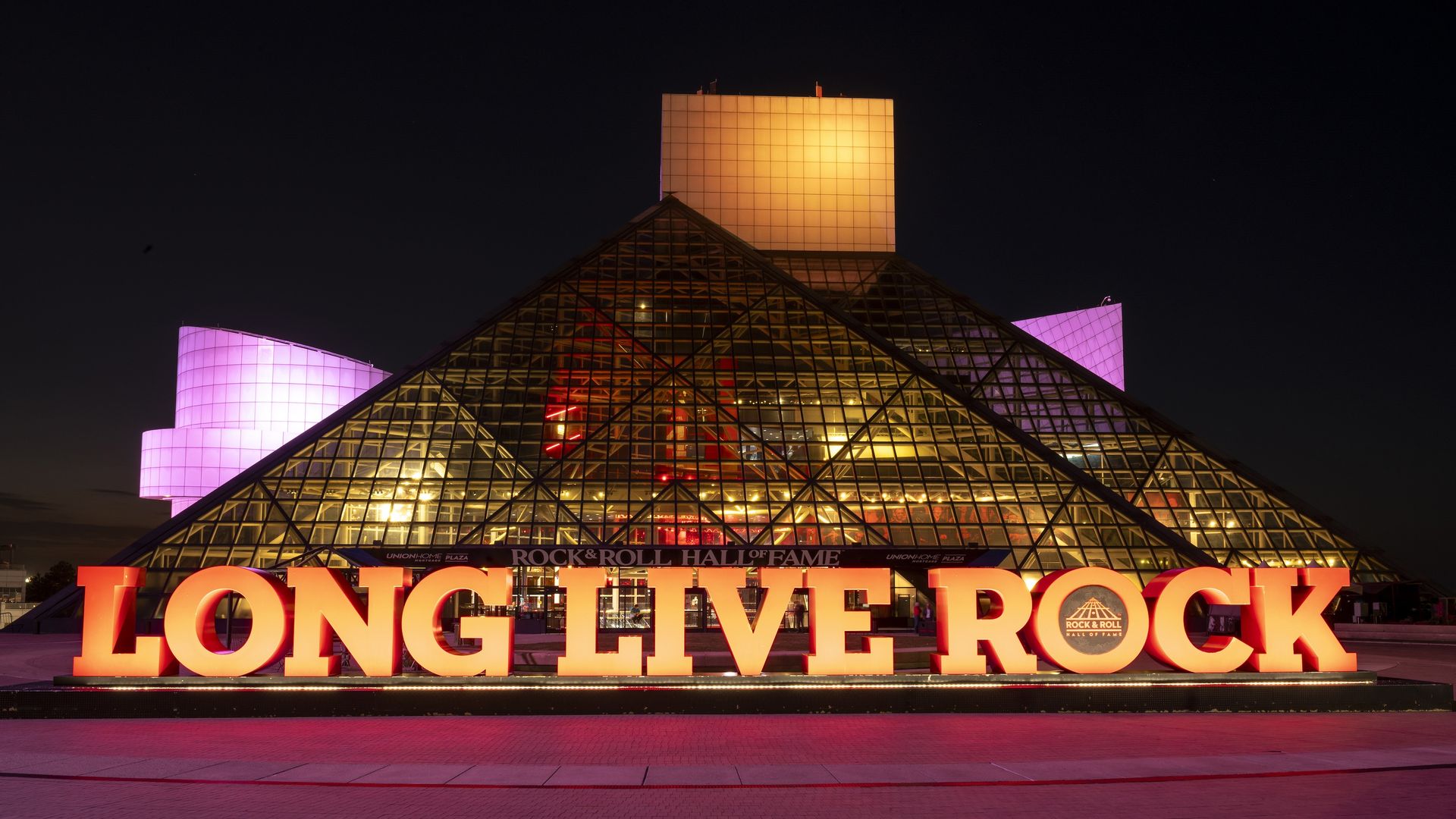 Night view of the Rock & Roll Hall of Fame with large illuminated orange letters spelling "LONG LIVE ROCK" in front of the pyramid-shaped glass building lit with pink and yellow lights.