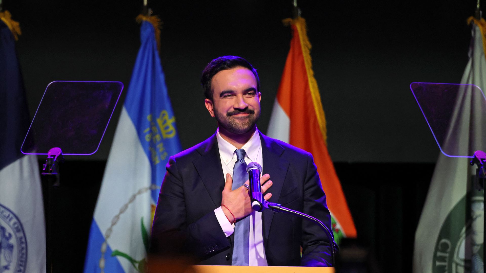 A dark-haired, bearded New York City Mayoral candidate Zohran Mamdani, wearing a navy jacket, white shirt and metallic blue tie, smiles as he puts his right hand on his chest while standing before a mic at a podium.