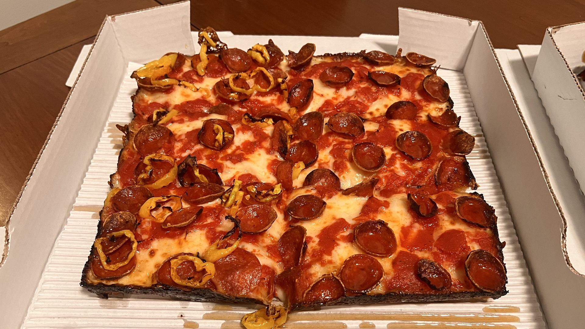 Square pepperoni and yellow pepper pizza with crispy edges served in a white cardboard box on a wooden table.