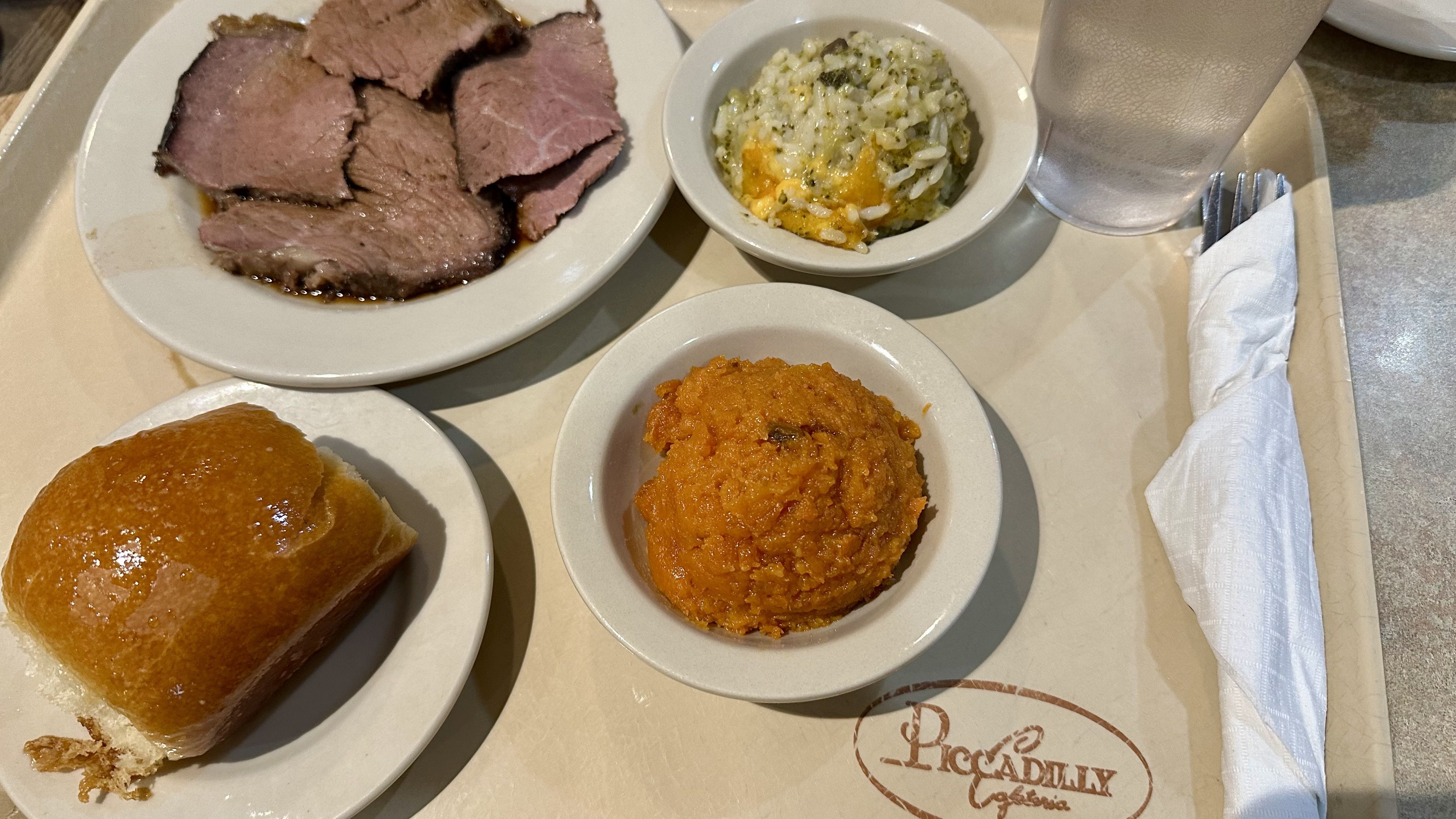 A meal tray with sliced roast beef in gravy, a bowl of cheesy rice, a bowl of carrot soufflé, a dinner roll, a glass of water, and wrapped silverware from Piccadilly Cafeteria.