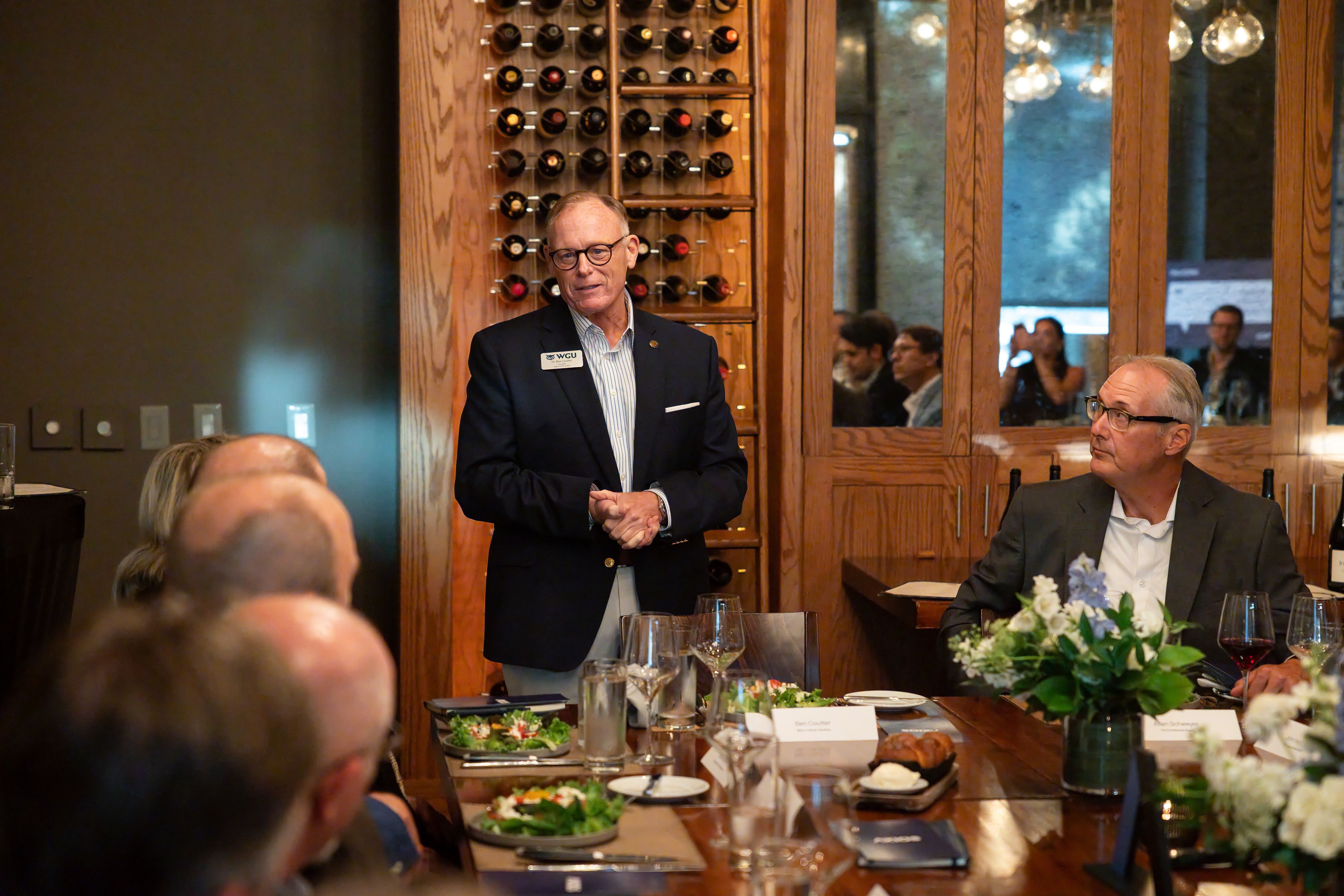 An older man in a dark blazer and glasses stands at a dining table giving a talk, with a wooden wine rack behind him; seated guests and a floral centerpiece are visible.