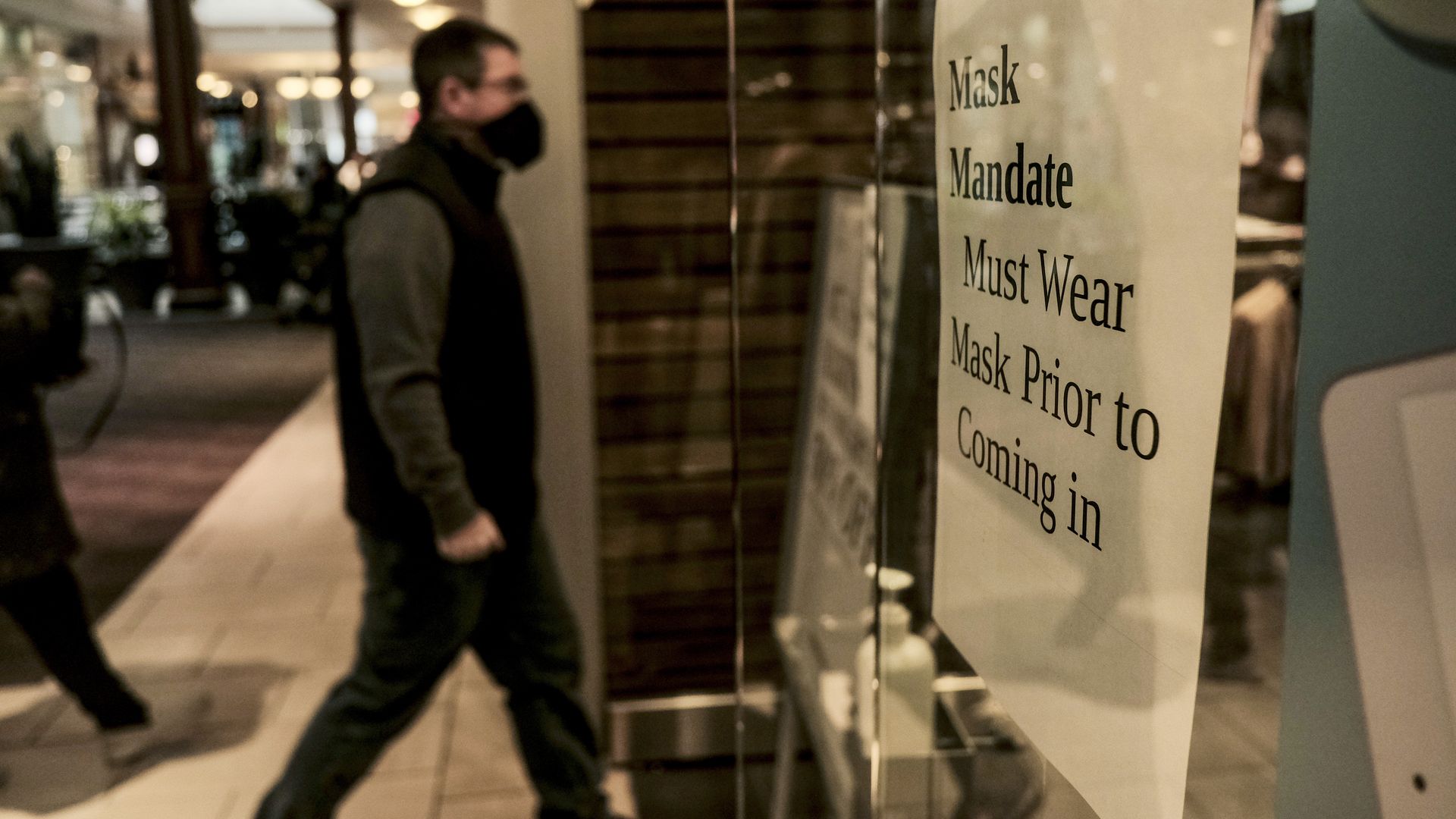 A man walks into a store with a sign on the window reading, "Mask Mandate Must Wear Mask Prior To Coming In."