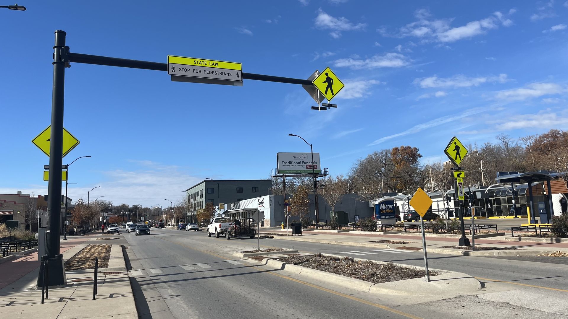 Urban street scene with pedestrian crossing signs in bright yellow, a state law sign instructing "STOP FOR PEDESTRIANS," clear blue sky, cars on road, and a Mister Car Wash sign.