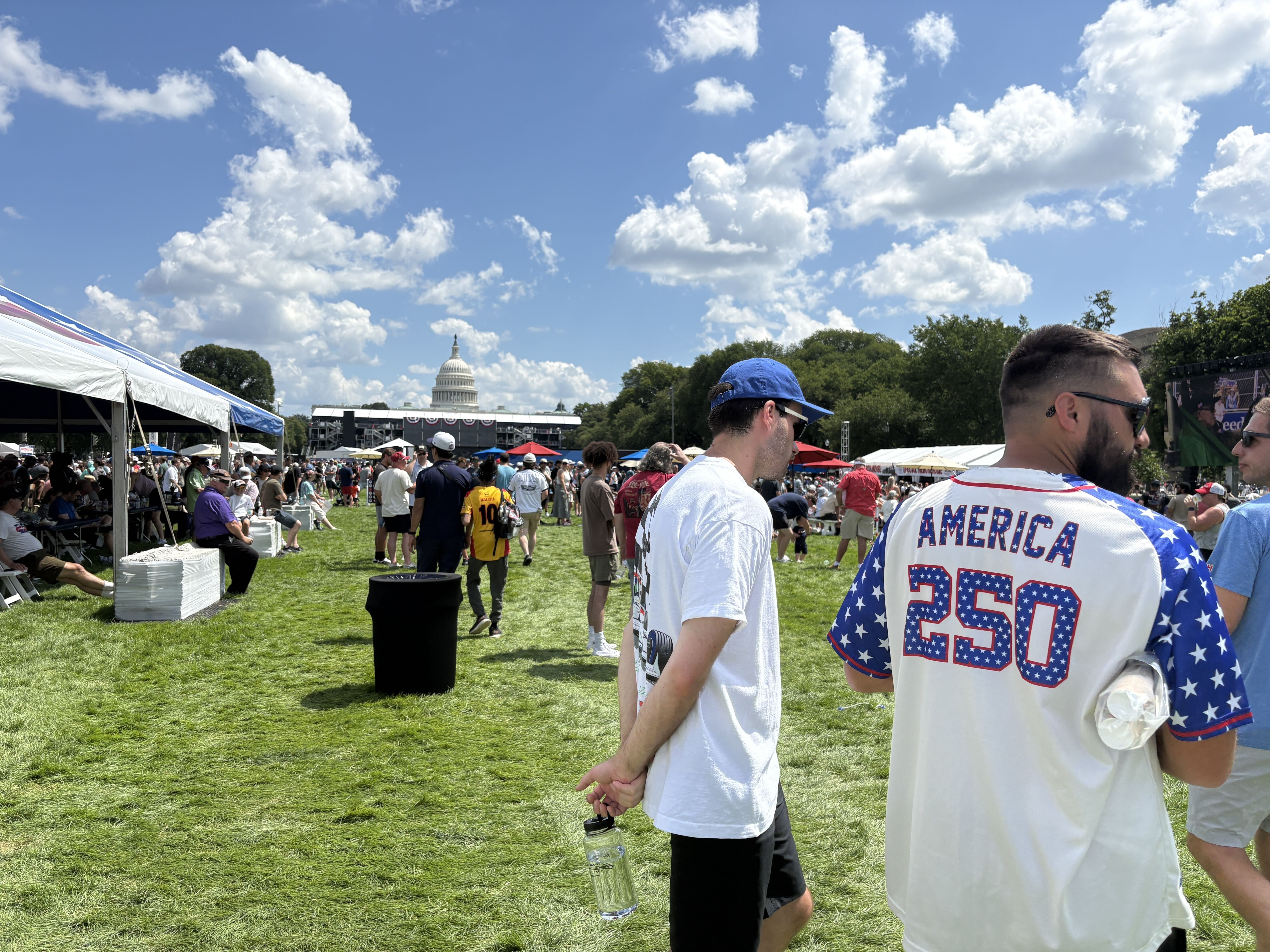 Wide outdoor festival on a bright day with white tents and a crowd on green grass; distant U.S. Capitol dome visible under a blue sky with fluffy clouds; attendees wear patriotic red, white, and blue.