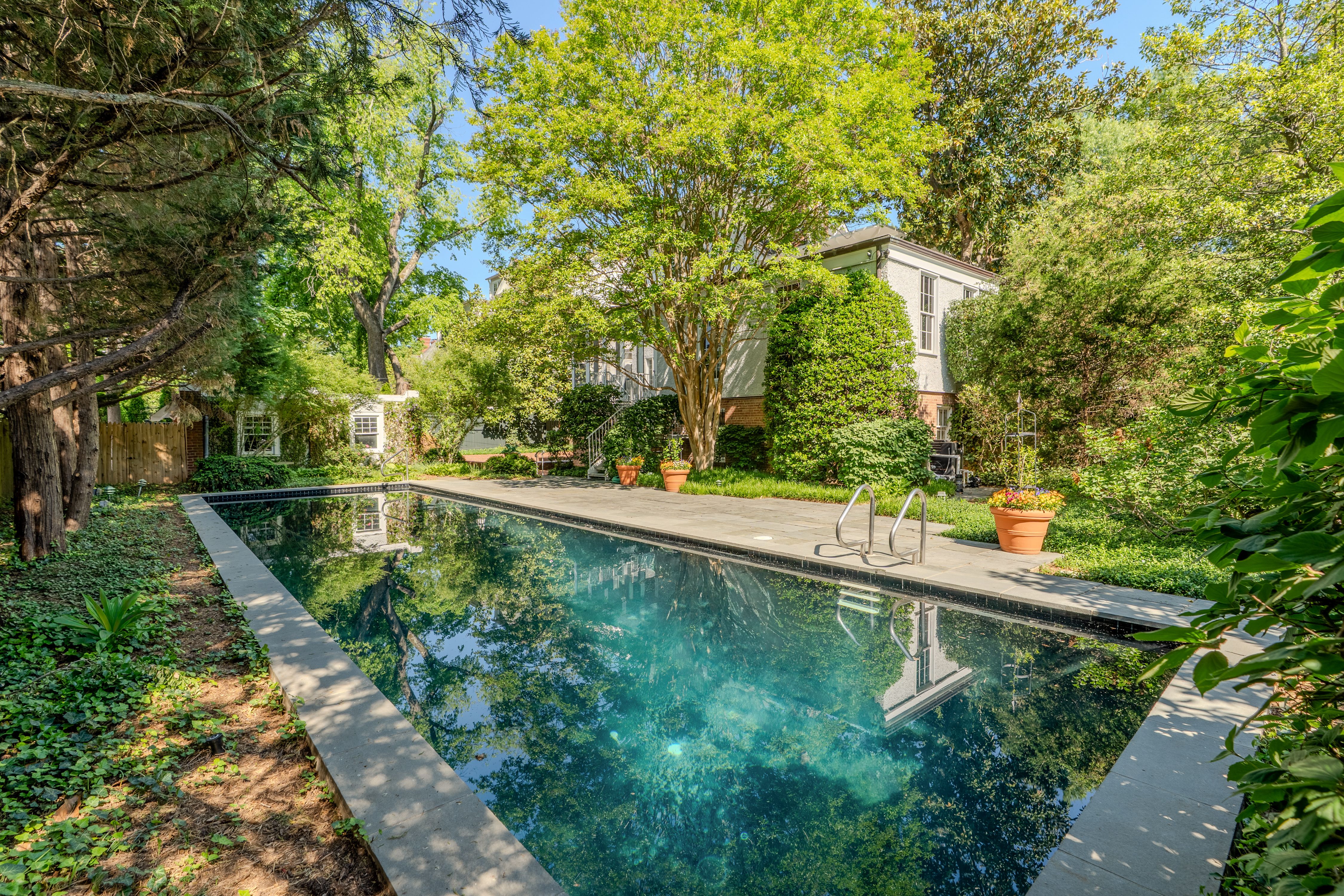 Sunlit backyard with a rectangular pool and concrete deck, surrounded by lush trees and hedges. A gray house with brick base sits behind, with potted plants by the pool and a blue sky.