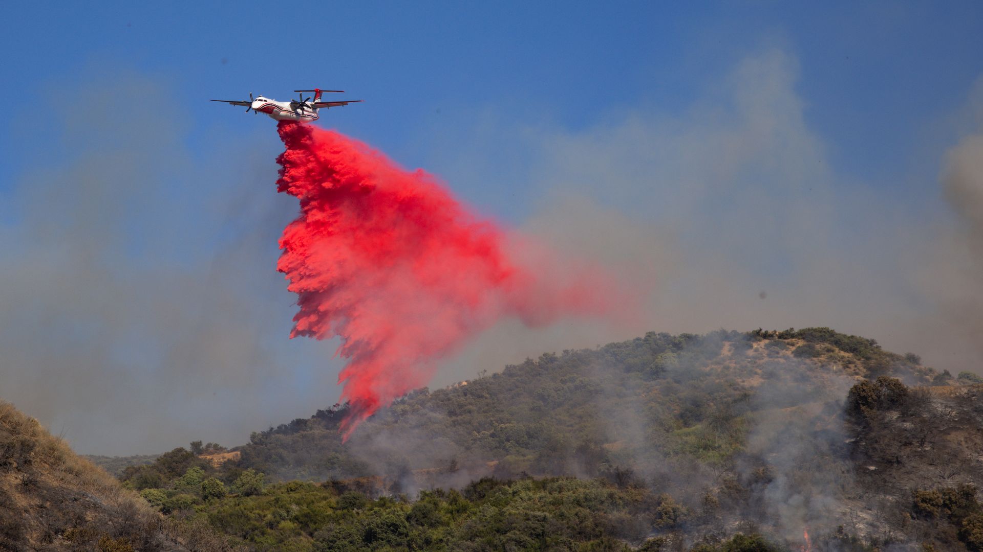 A Cal Fire plane drops a cloud of bright red fire retardant on hilly green forest with smoke rising and a small blaze.